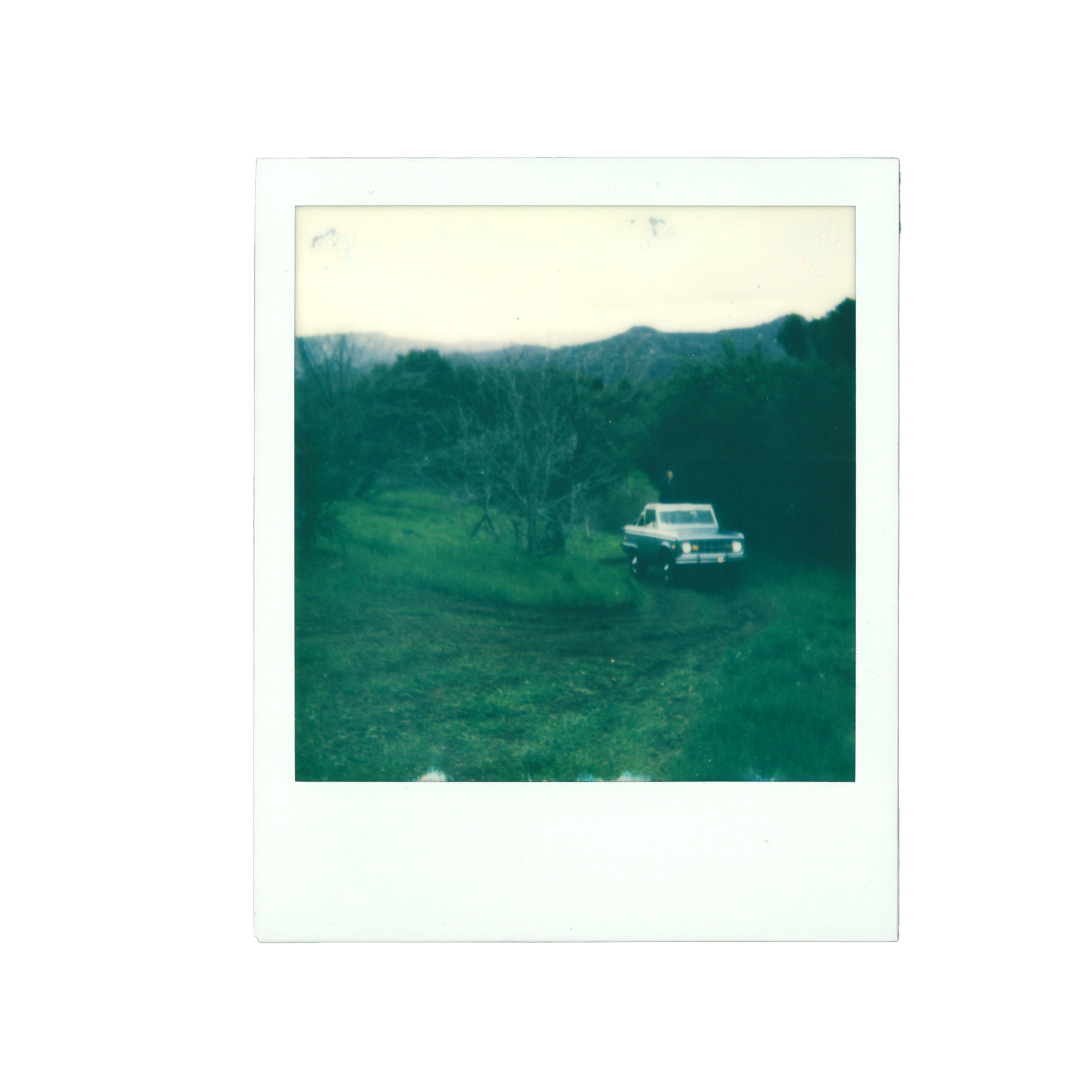 A vintage Polaroid picture showing a white and black SUV parked on a grassy field with trees and hills in the background during daytime.