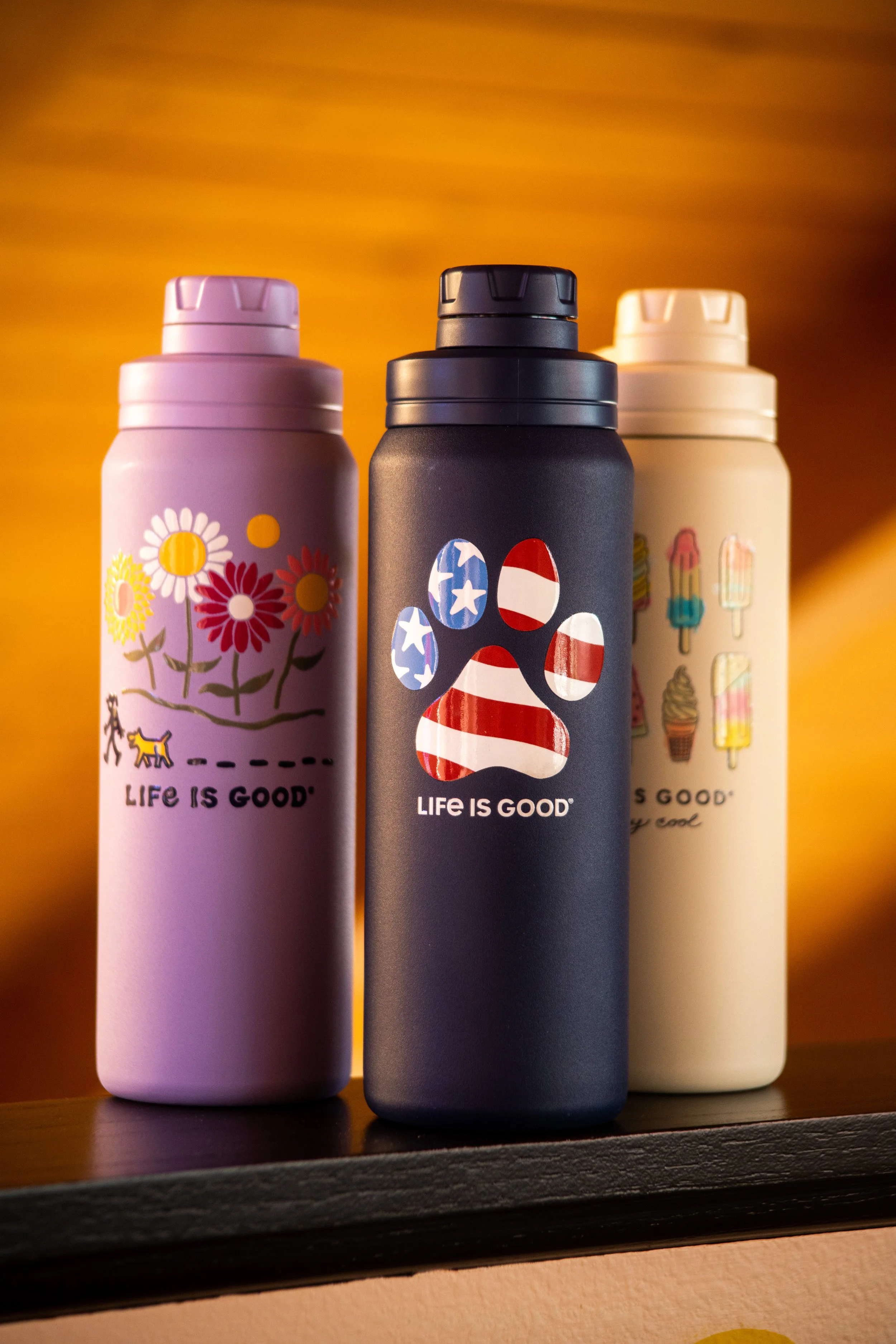 Three insulated bottles with colorful patterns and the phrase 'LIFE IS GOOD' on each, placed on a shelf against a warm, orange background.