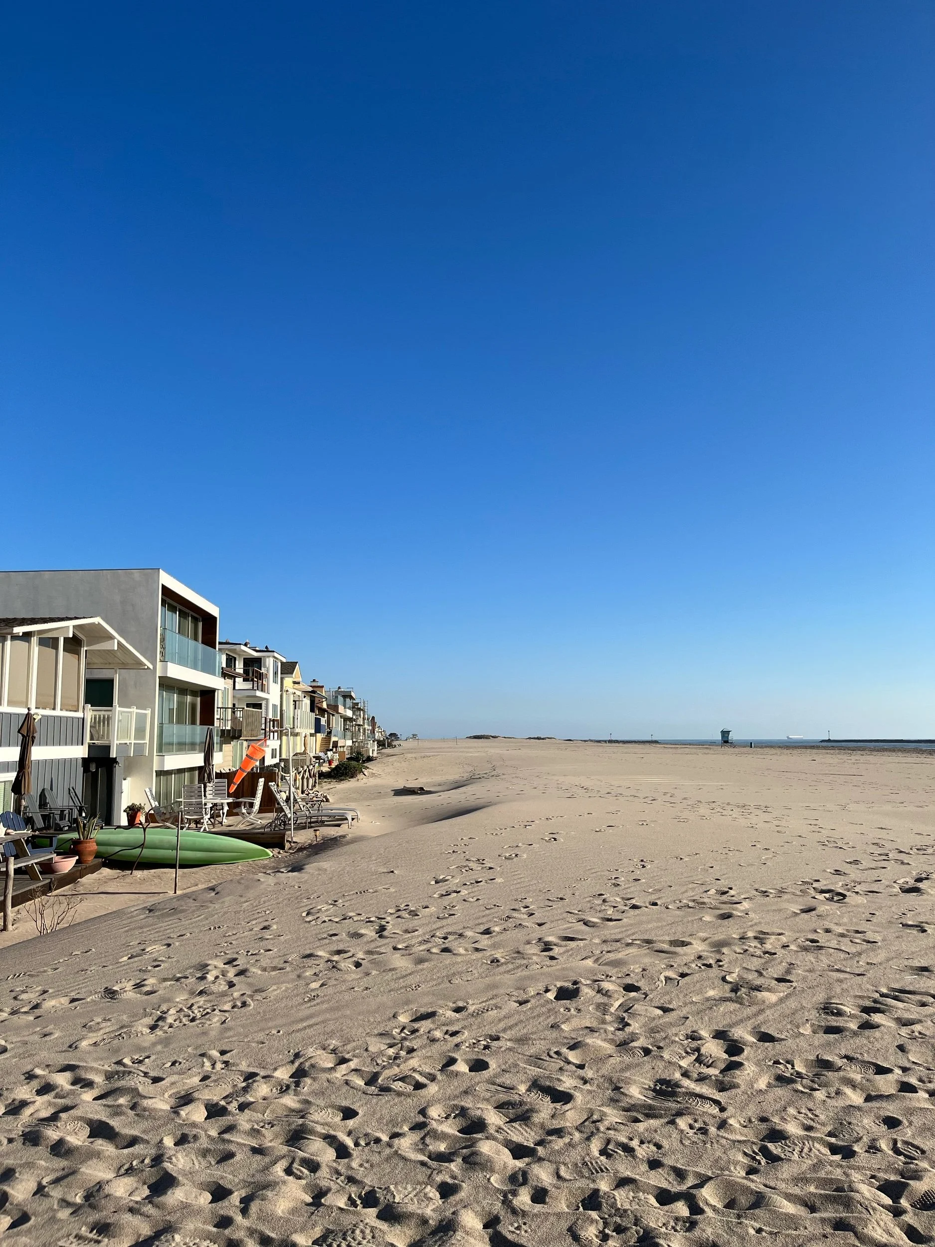 Empty sandy beach with footprints, a row of houses along the shoreline, a green kayak, and a clear blue sky.