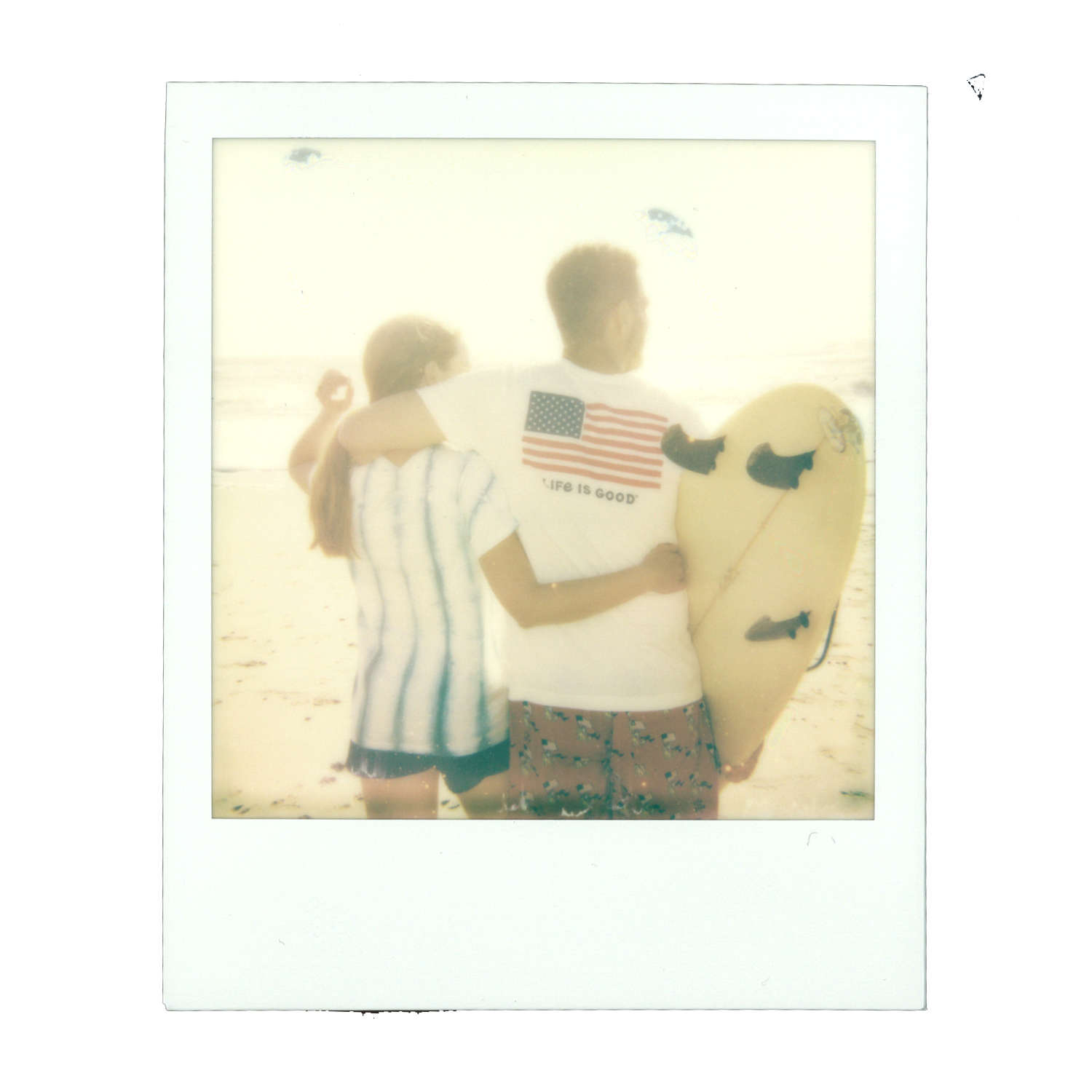 A young man and woman standing on a beach, with the man holding a surfboard and wearing a White T-shirt with an American flag and the phrase 'LIFE IS GOOD,' and the woman wearing a striped shirt, both facing away from the camera.