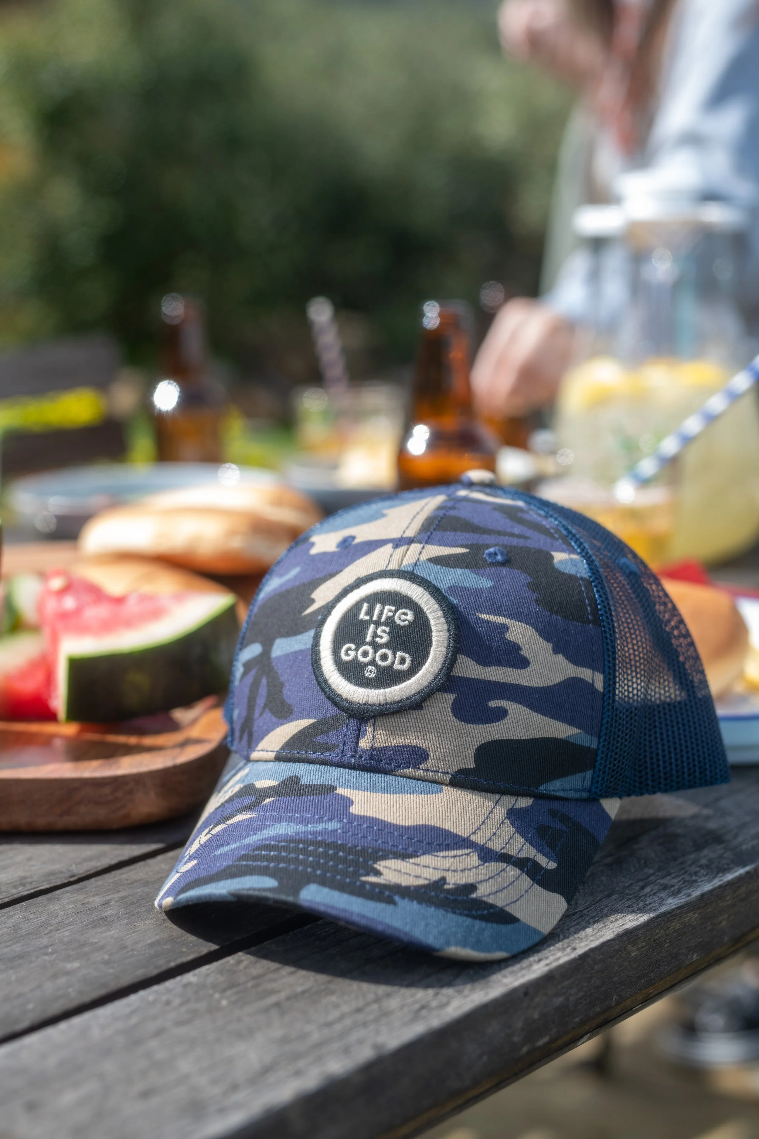 Camouflage baseball cap with the words "LIFE IS GOOD" embroidered on the front, placed on a wooden table outdoors with food and drinks in the background.
