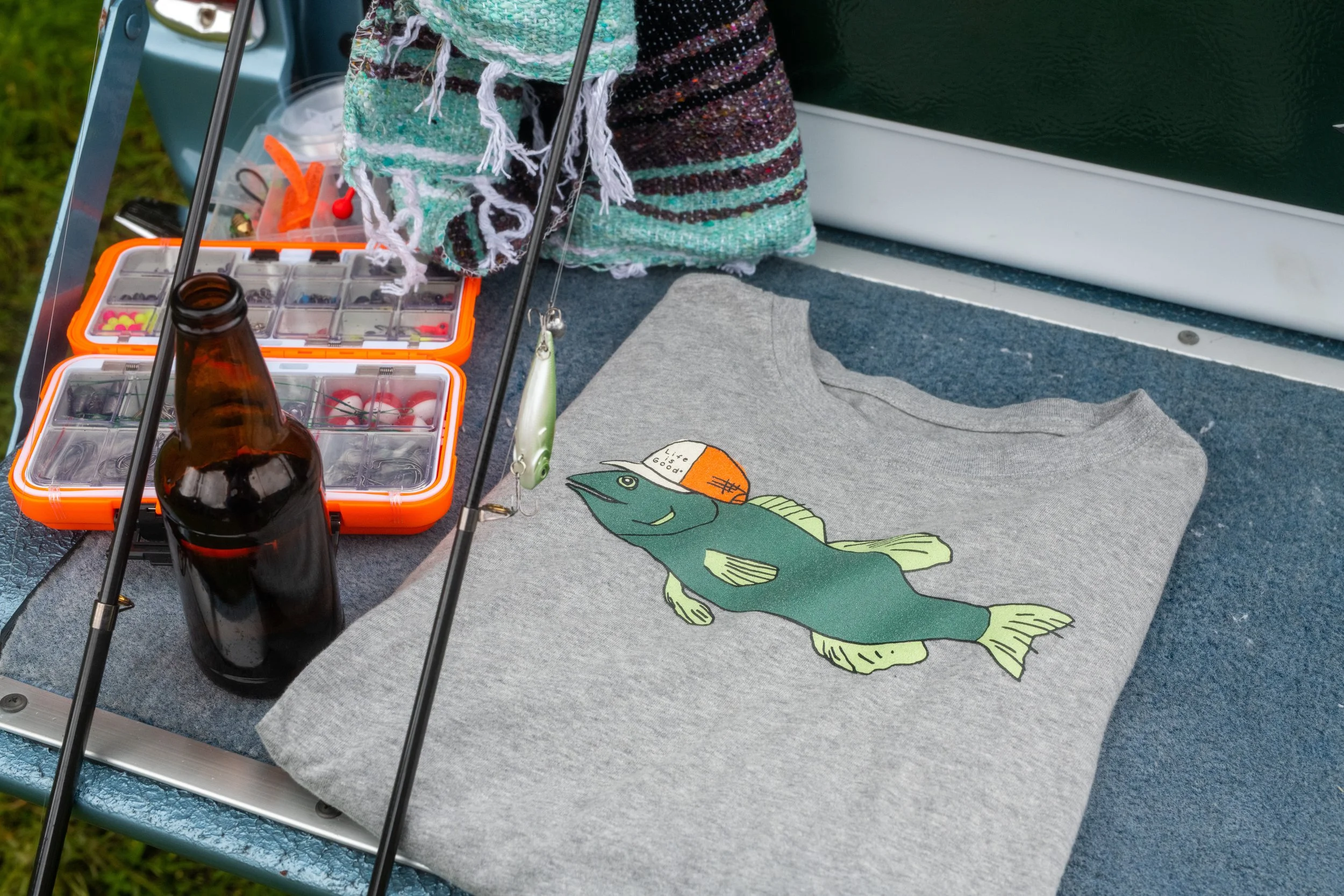 A gray T-shirt featuring a large fish graphic on it, with a small orange cap on the fish's head with the text 'Lax like a good' written on it. The shirt is laid out on a blue surface next to a fishing rod, a pattern cloth, an orange tackle box with f