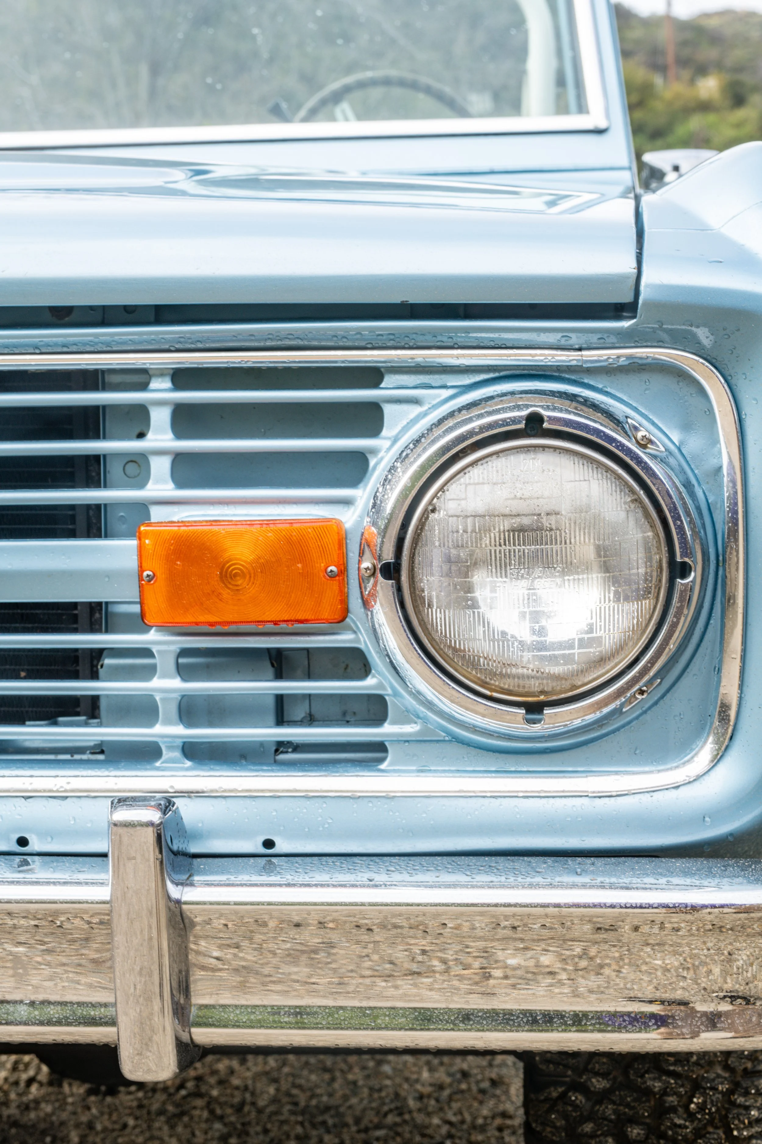 Close-up of the front of a vintage light blue car, showing the round headlight, orange turn signal, silver grille, and bumper.