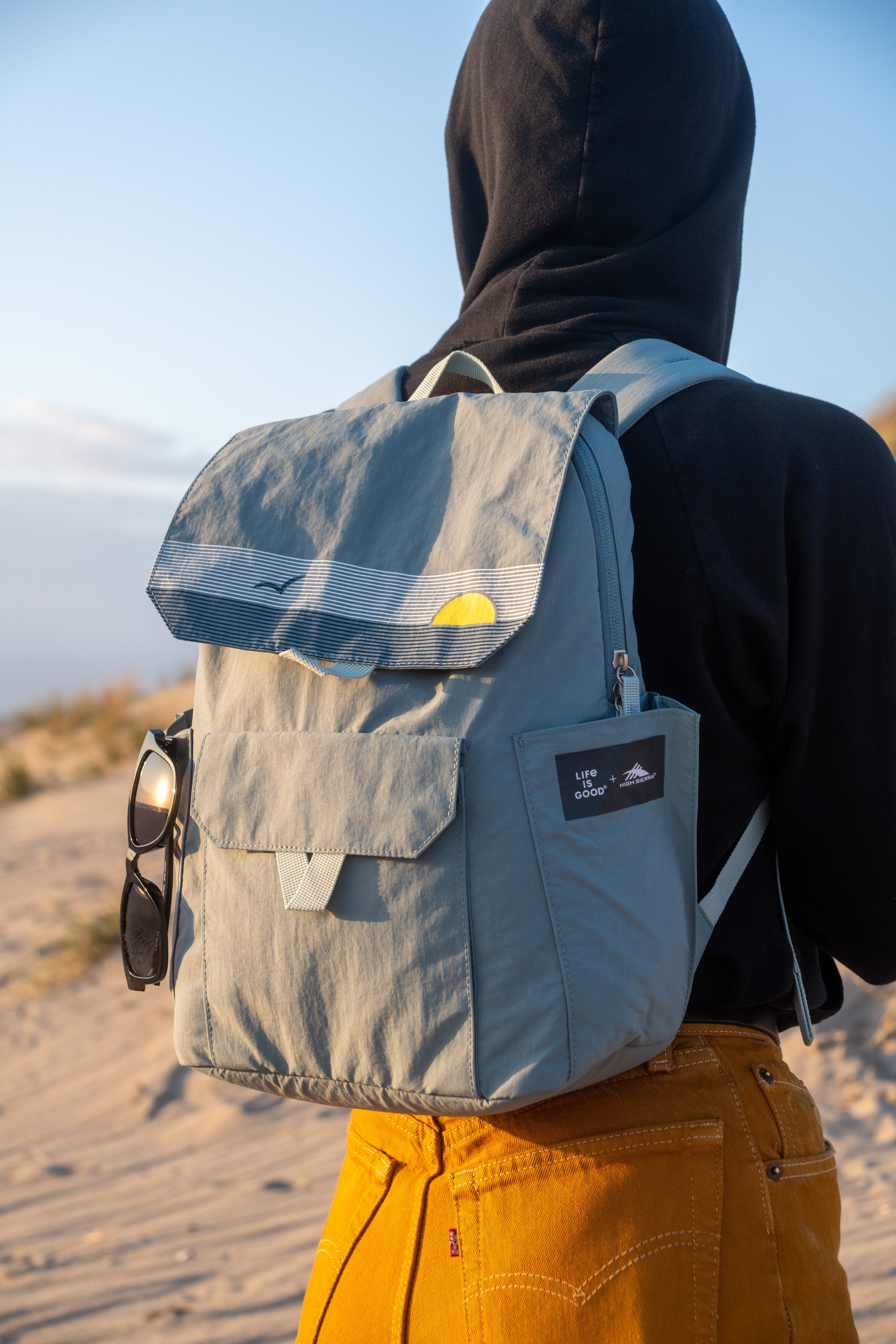 Person wearing black hoodie with yellow pants, carrying a gray backpack with a pair of sunglasses hanging from the front pocket, standing on a sandy outdoor landscape during daylight.