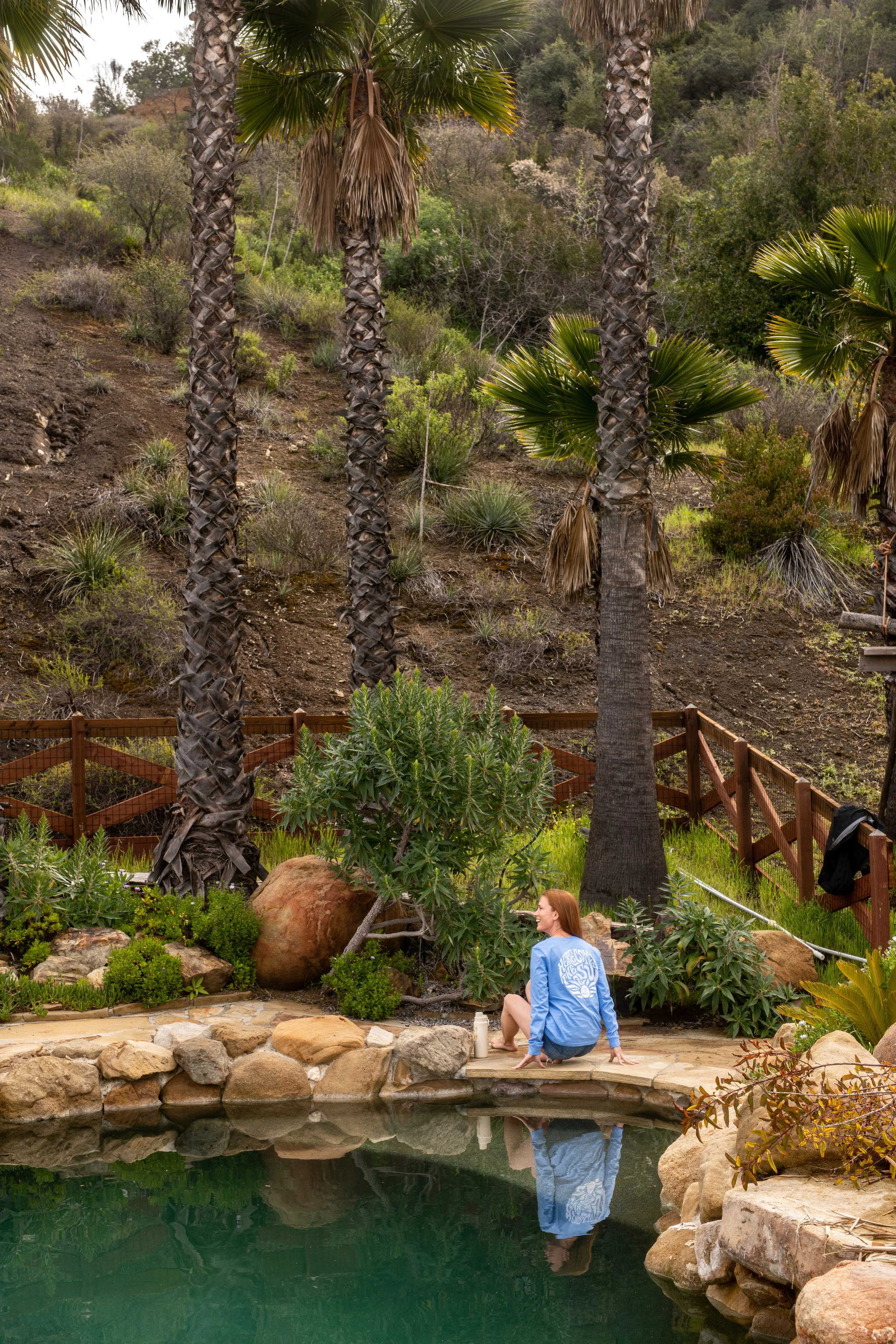 A person sitting on the edge of a small pond surrounded by rocks, looking and smiling at their reflection. The scene has lush green plants, palm trees, and a wooden fence in the background, set in a garden or natural landscape.