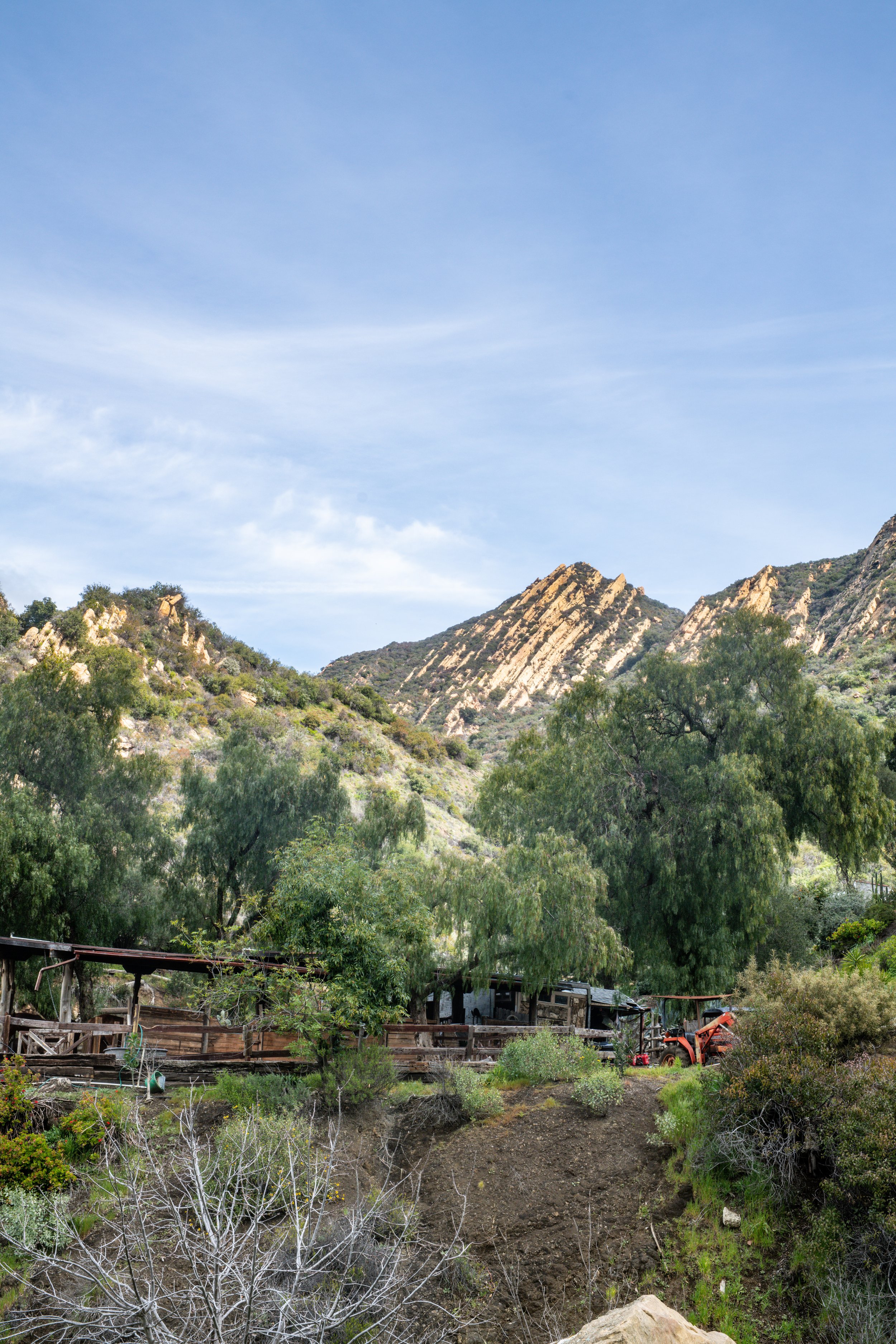 A scenic view of a mountainous landscape with rocky ridges and green trees, a clear blue sky, and a small rustic farm with trees, a wooden fence, and farm equipment in the foreground.