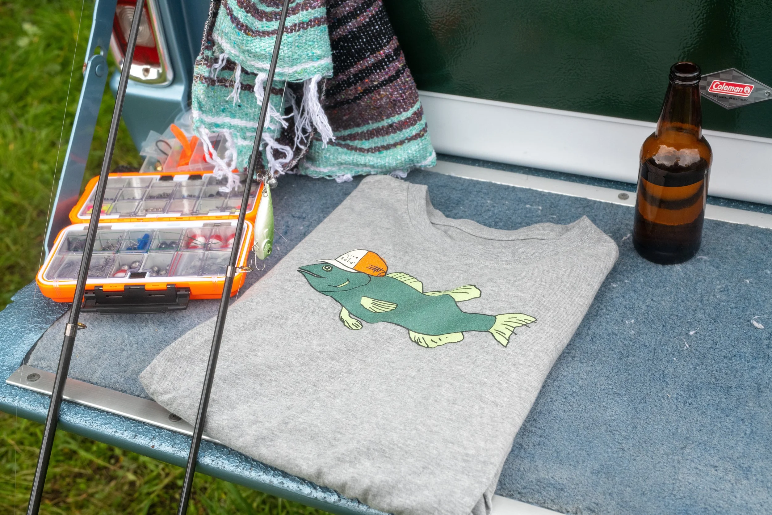 A gray T-shirt with a fish graphic, a fishing lure, a fishing tackle box, a brown beer bottle, and cloth towels with a colorful stripe pattern on a boat deck, with grass and water in the background.