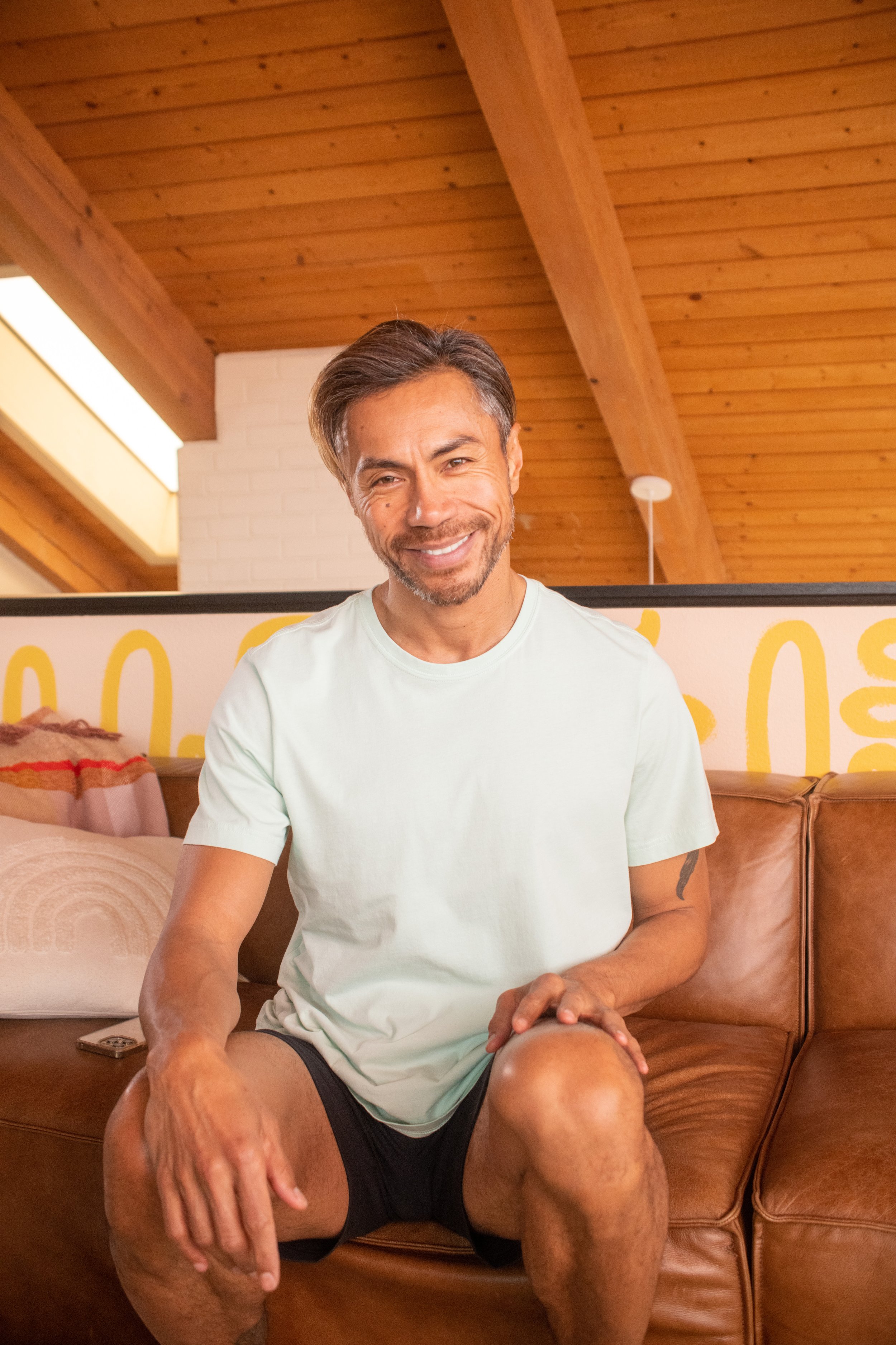 A smiling middle-aged man with short, styled hair and a beard, sitting on a brown leather couch in a room with a wooden ceiling and a white brick wall behind him.