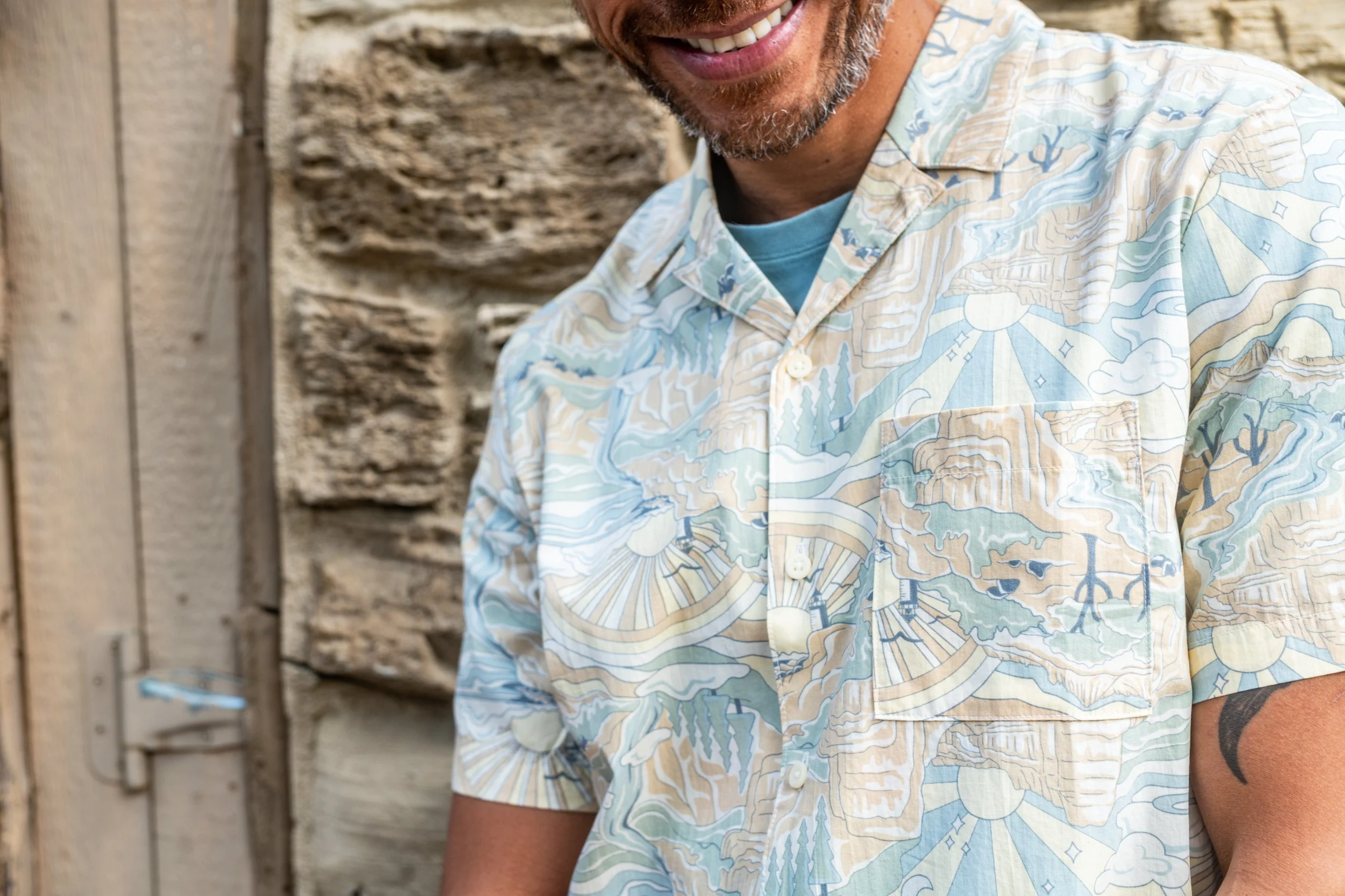 Close-up of a smiling man in a patterned short-sleeve shirt, standing outdoors near a stone wall.