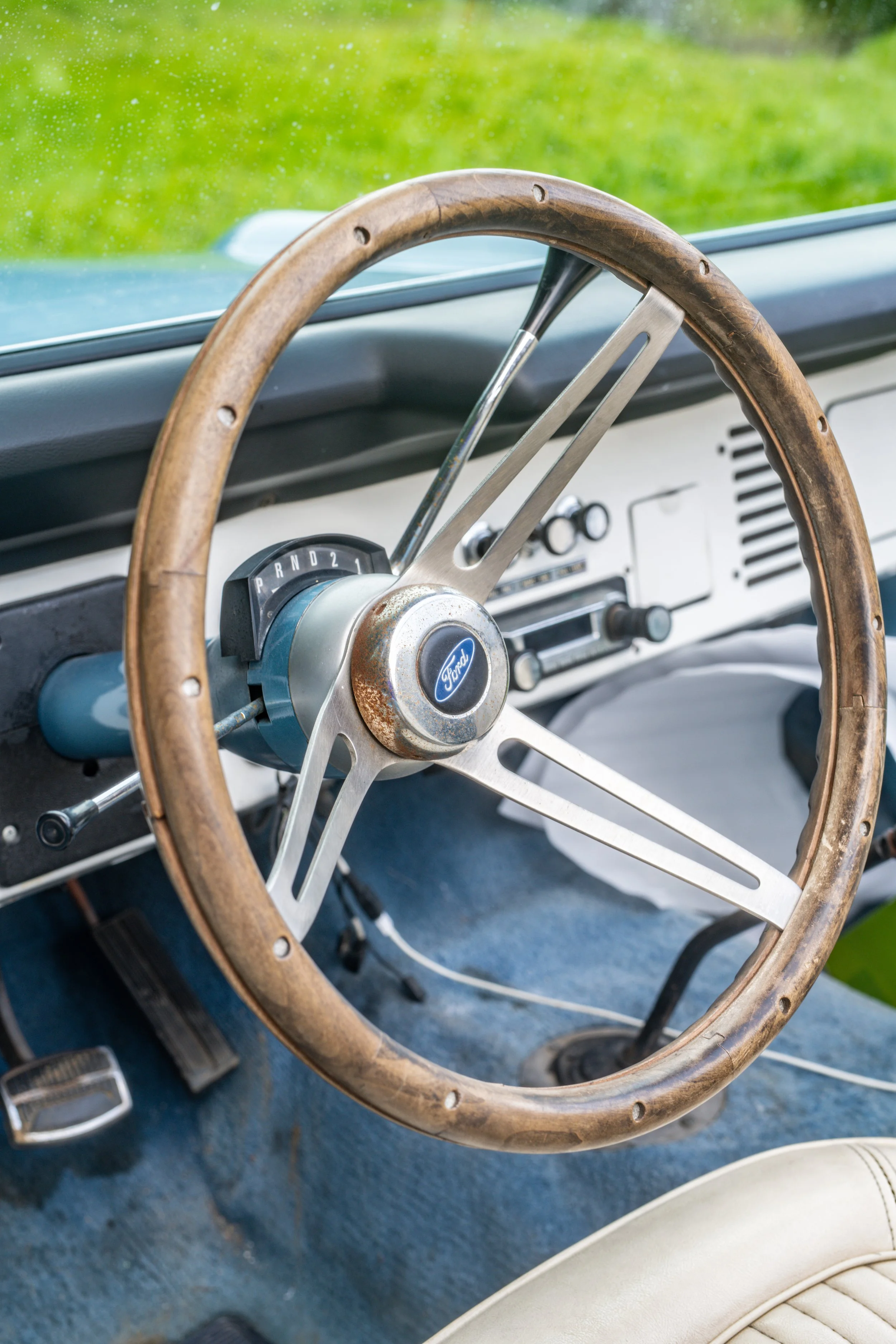 Close-up of a vintage car interior featuring a wooden steering wheel with metal spokes, a Ford logo at the center, and an automatic gear shift with the settings PRND21, with a view of the windshield showing greenery outside.