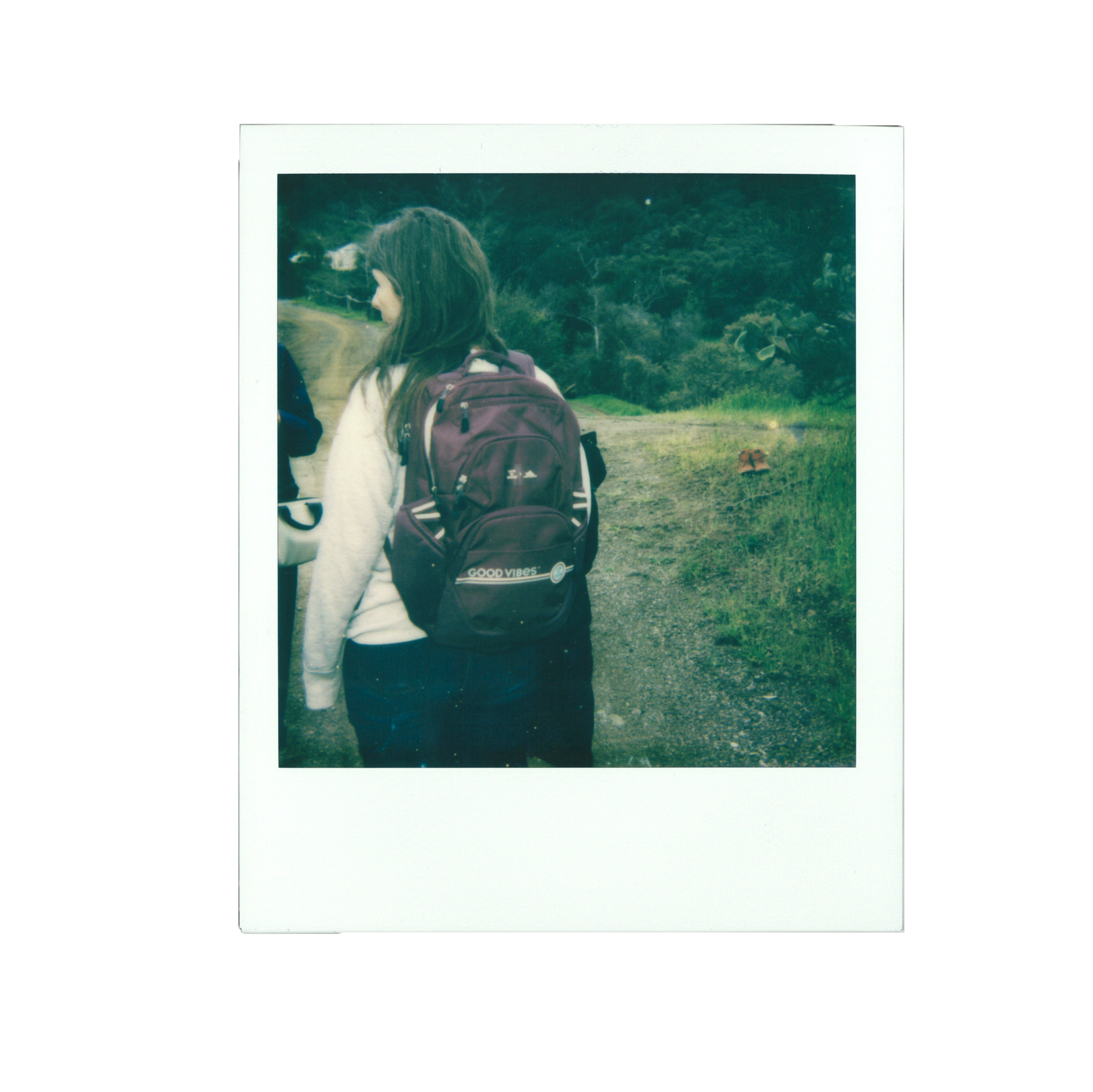 A young woman with long brown hair, wearing a white sweater and carrying a maroon backpack, is standing on a dirt path surrounded by greenery.