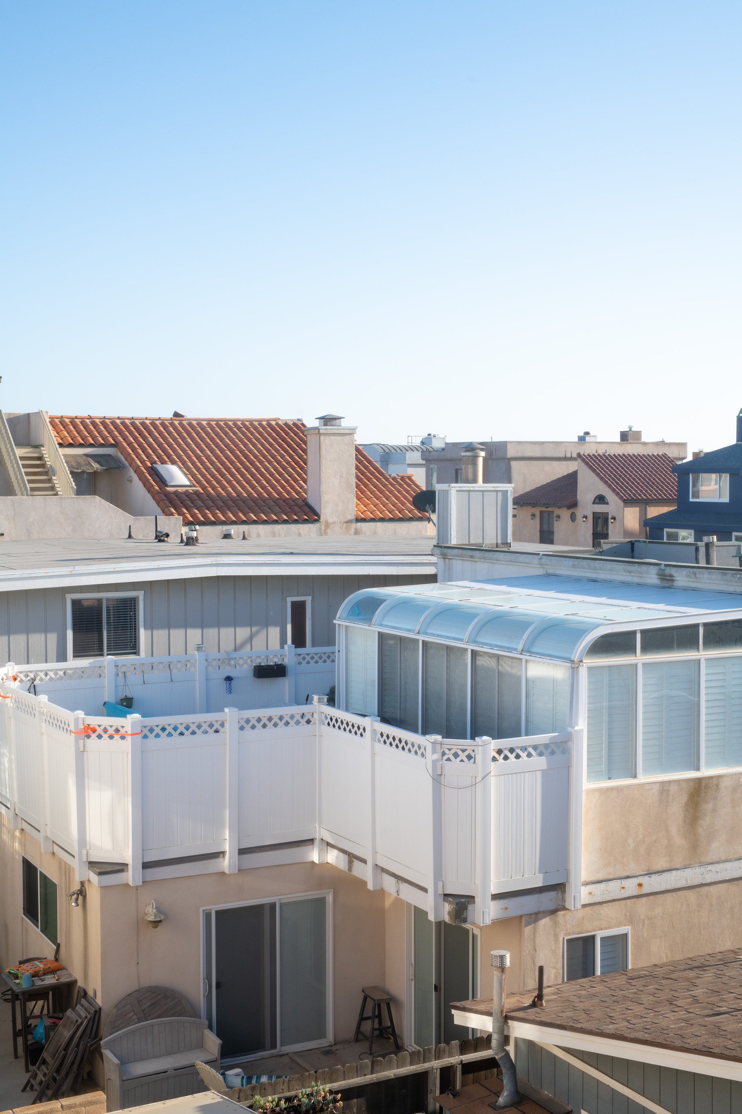 View of a neighboring residential area with rooftops, including shingles and tiles, and a backyard patio with a white fence and a small enclosed glass room or sunroom.