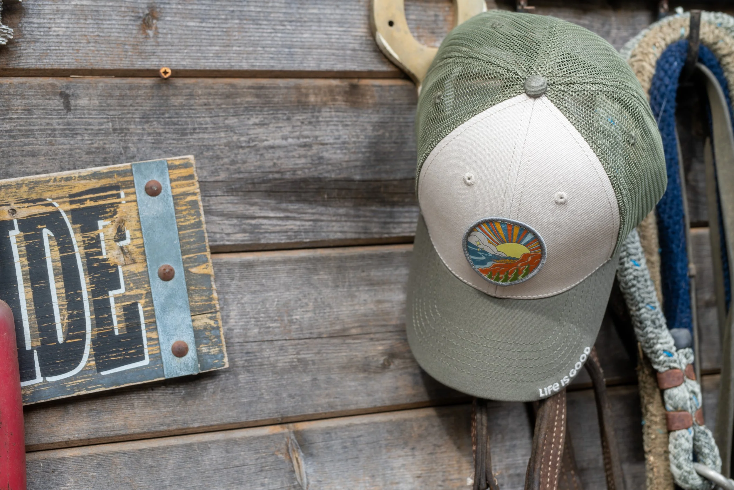 A beige and green trucker hat with a circular patch of a sunset over mountains and a river hanging on a weathered wooden wall, with a rope leash and a chain on the right side.