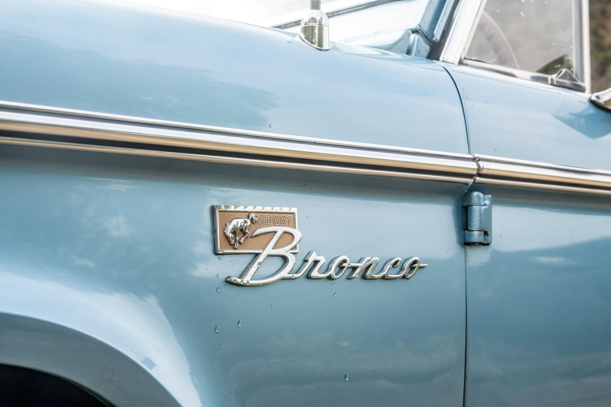 Close-up of the side of a light blue vintage Ford Bronco Sport vehicle, showing the Bronco emblem and badge with a horse graphic.