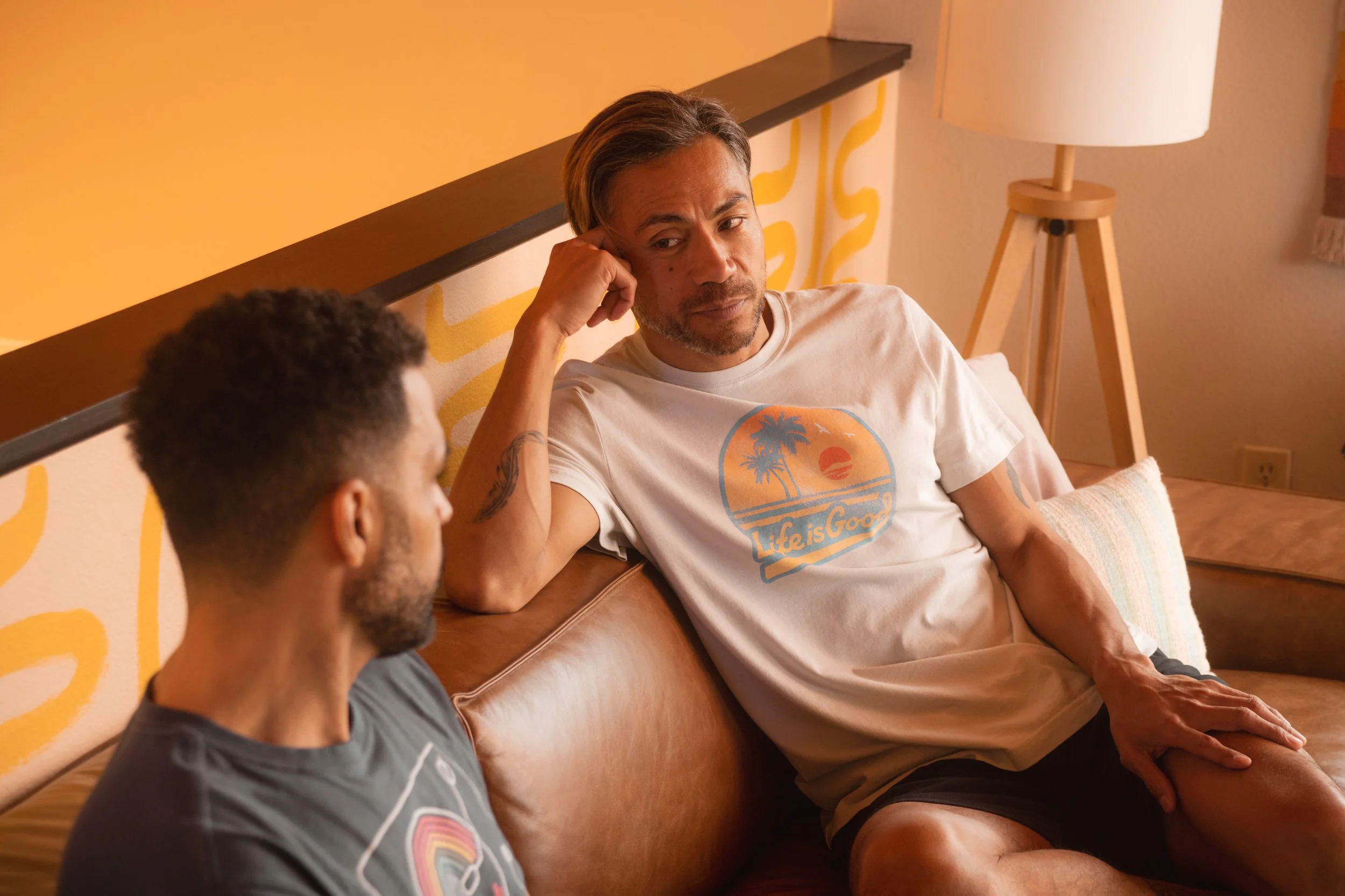 Two men sitting on a leather couch in a cozy room, one with his arm resting on the back of the couch, engaged in conversation. The man on the right has tattoos, wears a white t-shirt with a sunset and palm tree design that says 'Life is Good,' and ha
