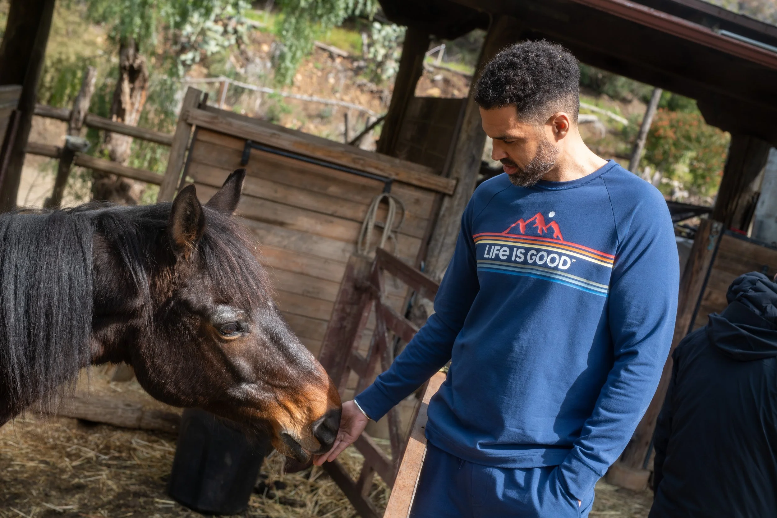 A man with curly hair and a beard in a blue sweatshirt that says 'LIFE IS GOOD' stands in a rustic barn, gently petting a brown horse's nose.