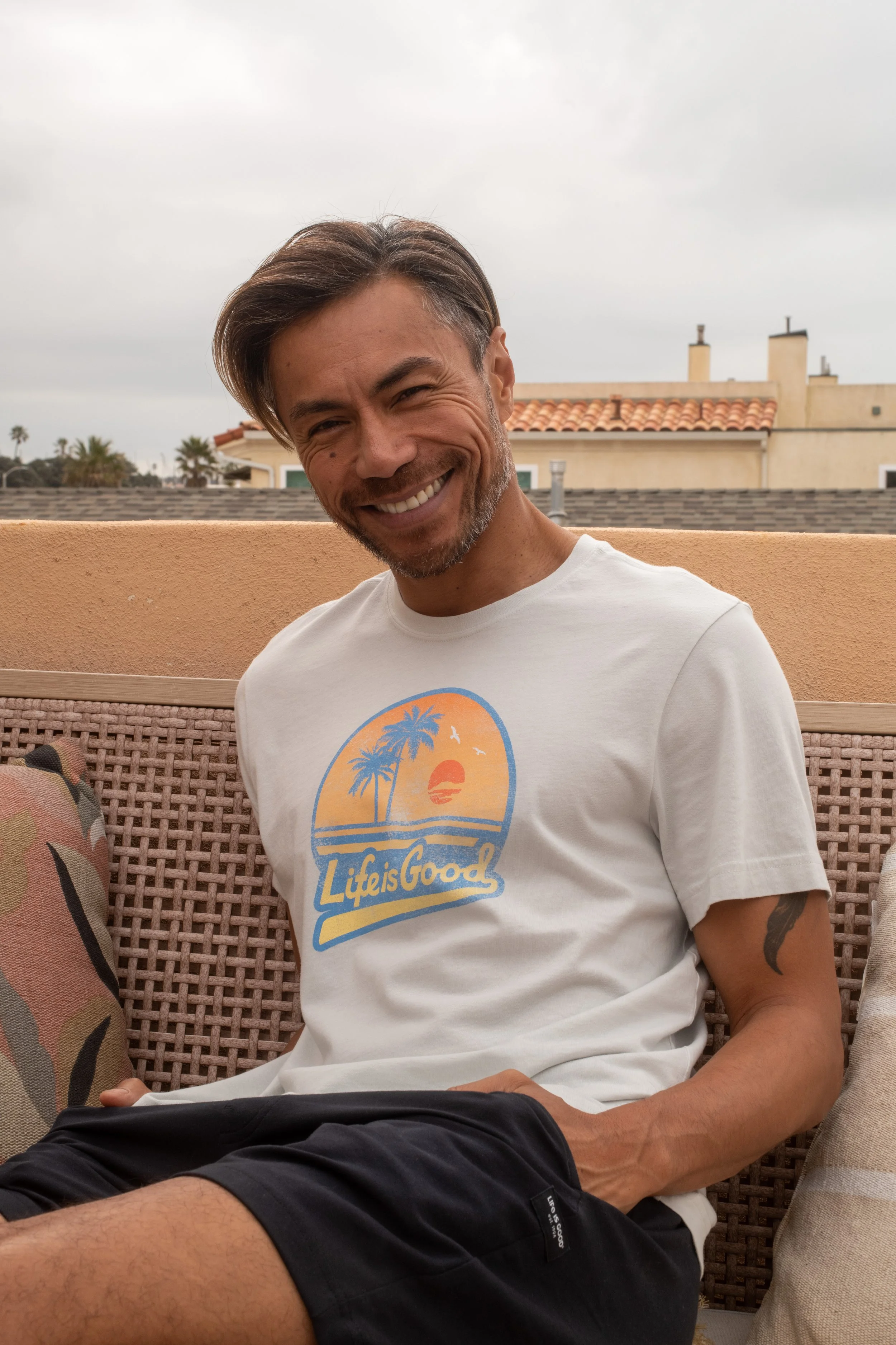 A smiling man sitting outdoors on a patio, wearing a white t-shirt with a sunset and palm trees graphic and the text "Life is Good."