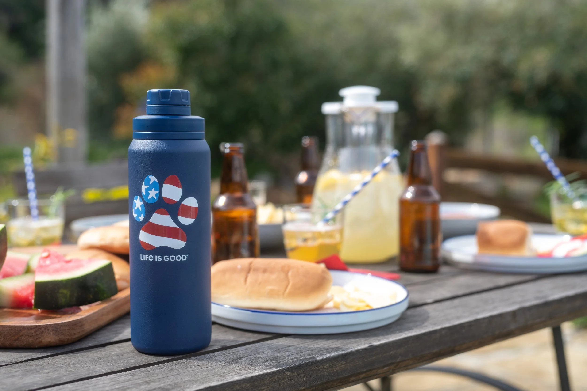 Outdoor picnic table set with plates of food, bottles, and a blue water bottle with a paw print and American flag design that says 'Life is Good'.