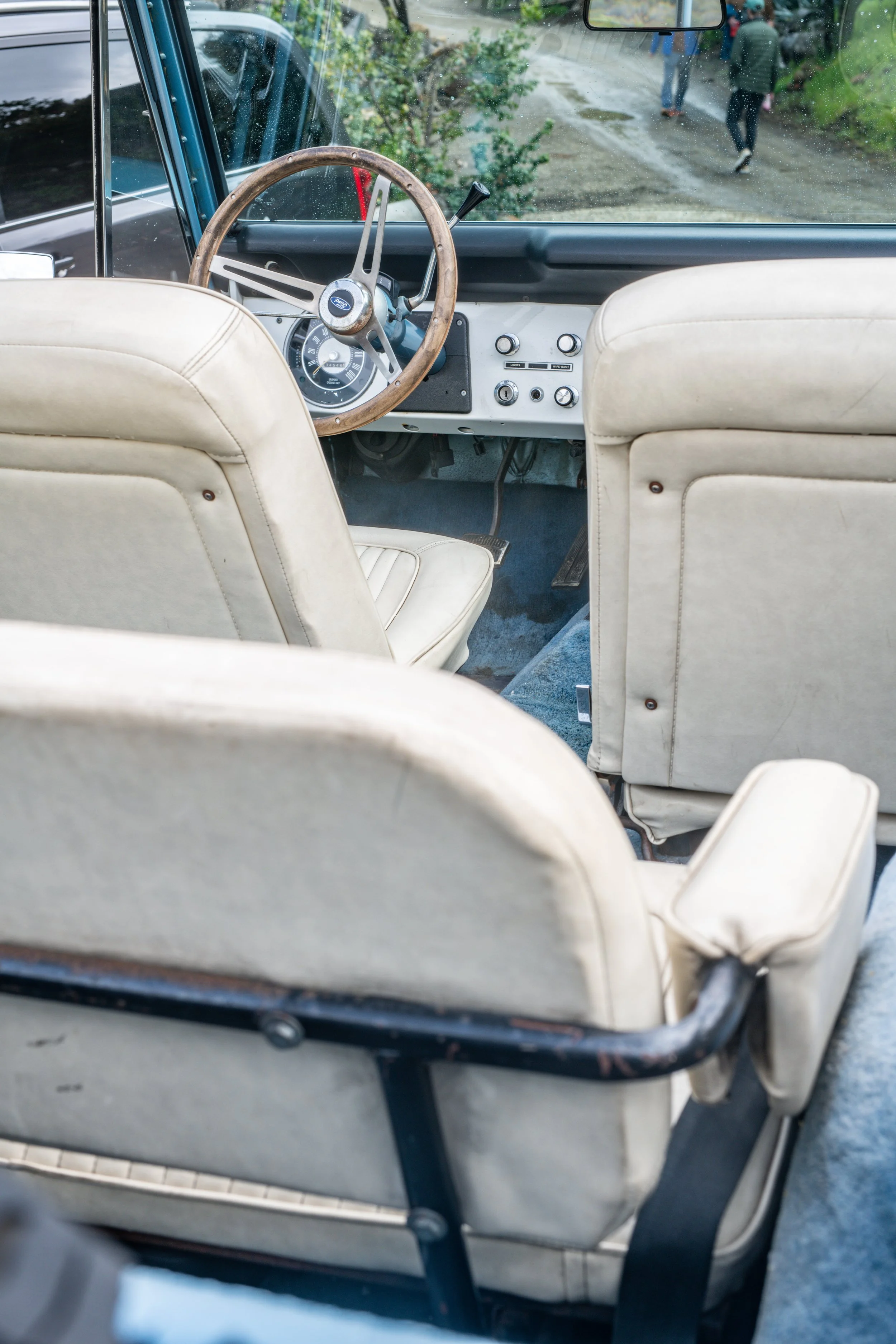 Interior view of an older vehicle with cream-colored seats, a wooden steering wheel, and a dashboard with vintage controls, parked on a dirt road with people walking nearby.