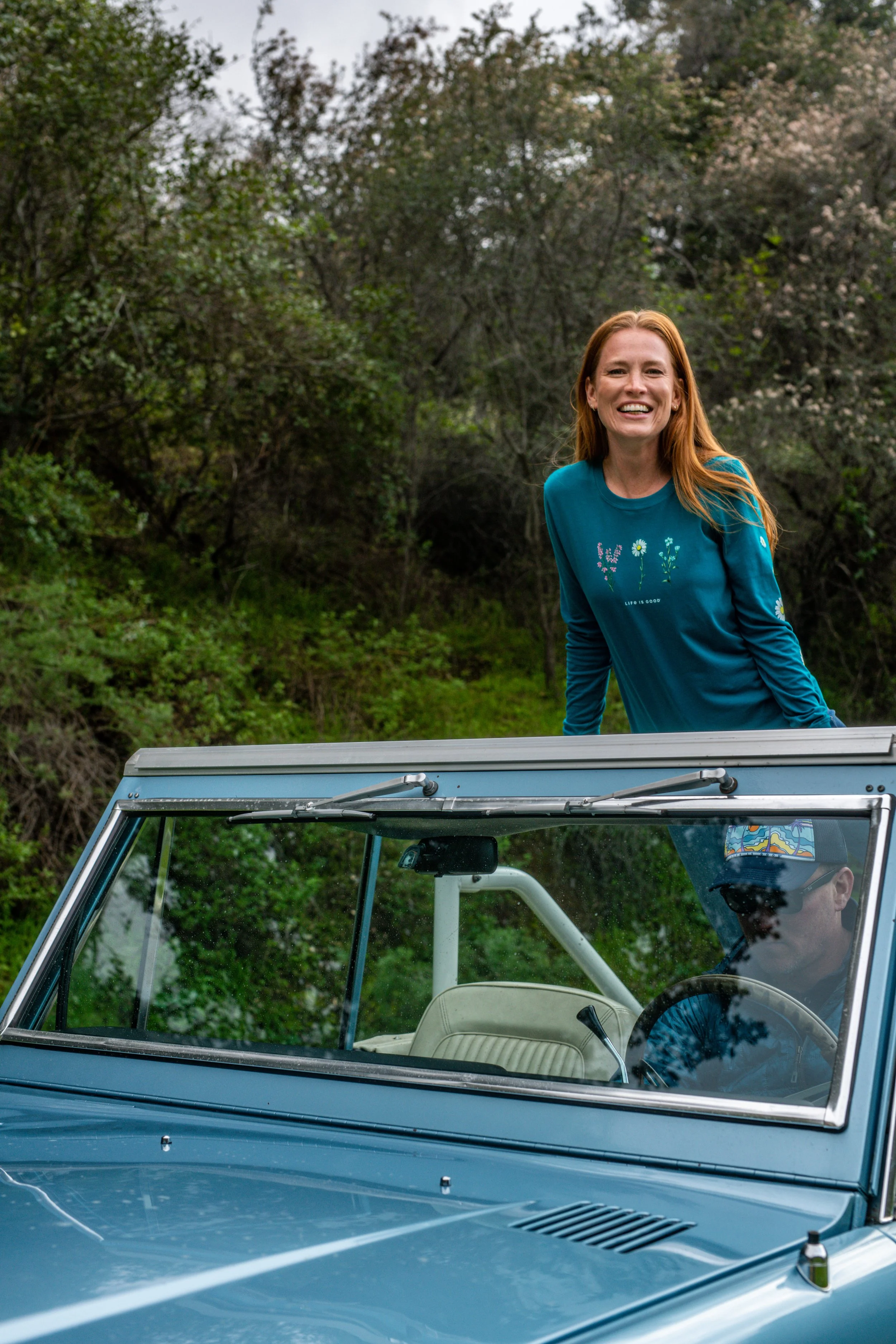 A woman with red hair and a teal long-sleeve shirt is smiling and leaning on a vintage blue convertible car, with a man driving inside, in a lush green outdoor setting.