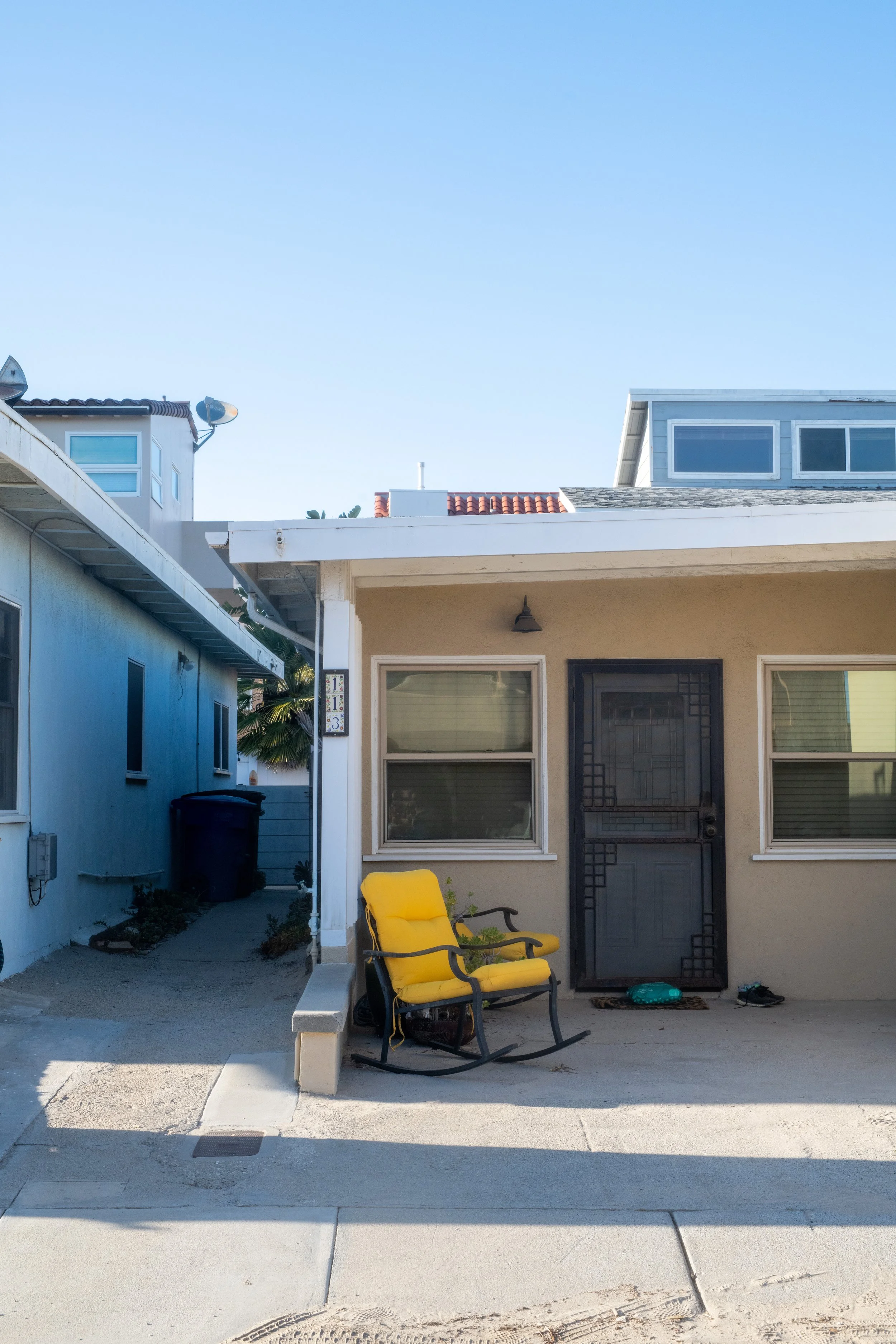 Front of a house with a yellow outdoor rocking chair, two windows, a black screen door, and a small pathway leading to the side yard.