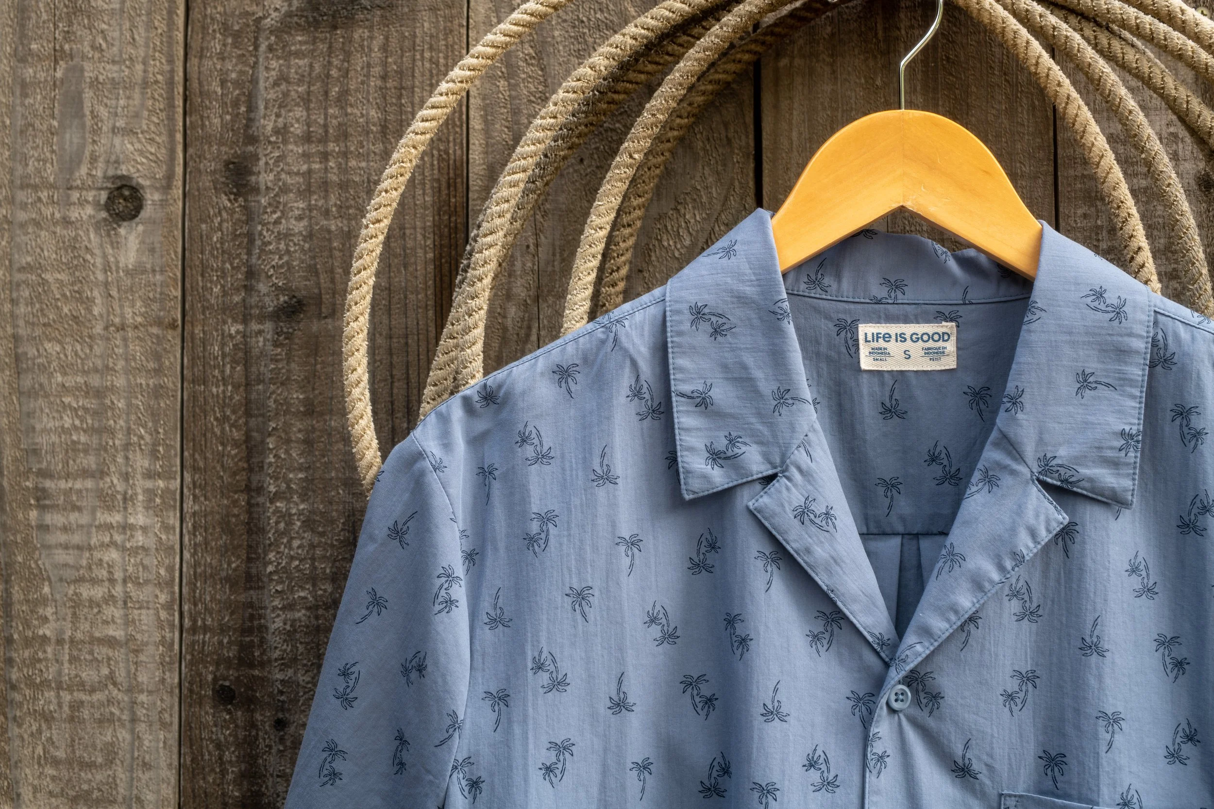 A light blue short-sleeve button-up shirt with a small palm tree pattern hanging on a wooden hanger in front of a rustic wooden wall with decorative rope accents.