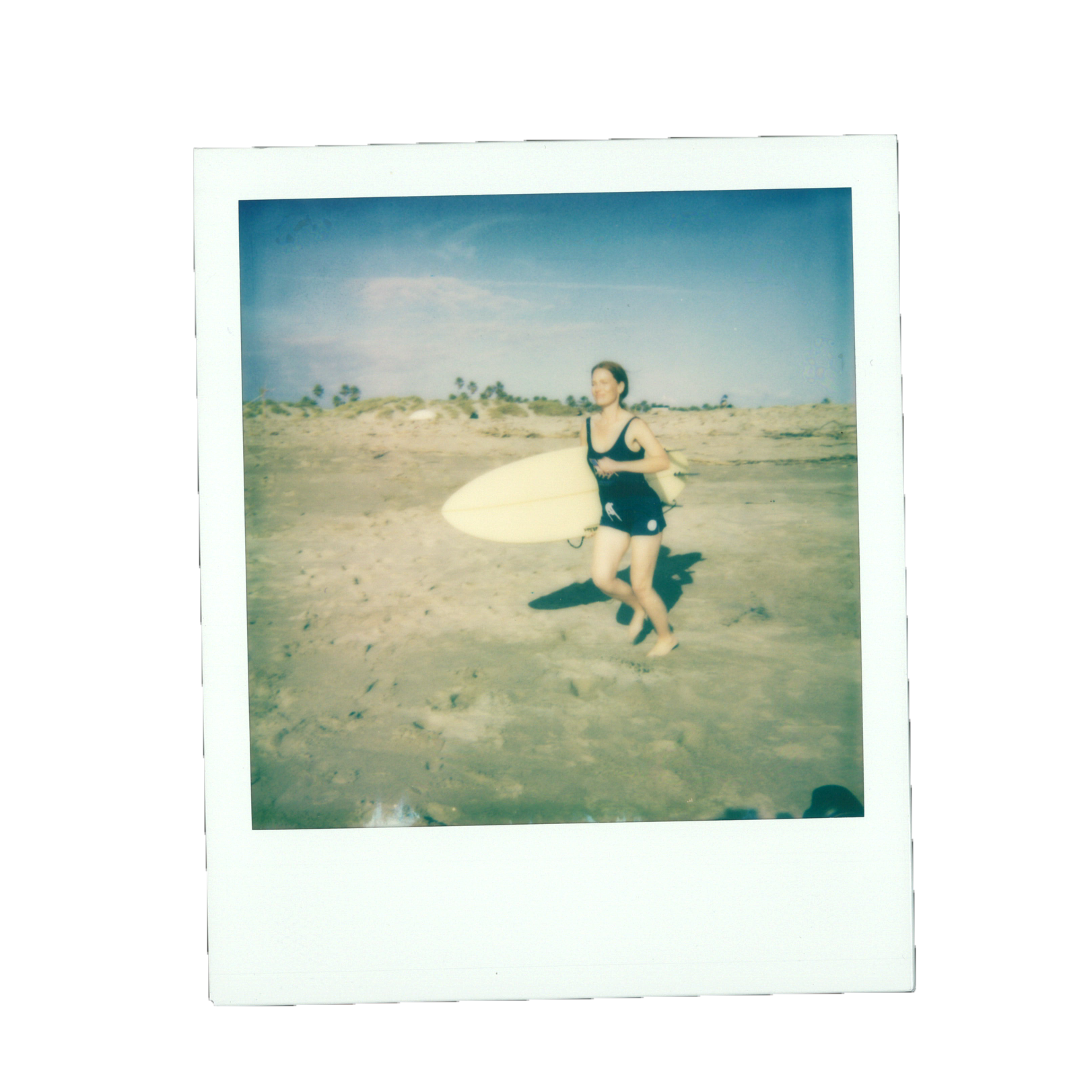 A woman in a black swimsuit holding a surfboard on a sandy beach with blue sky and palm trees in the background.
