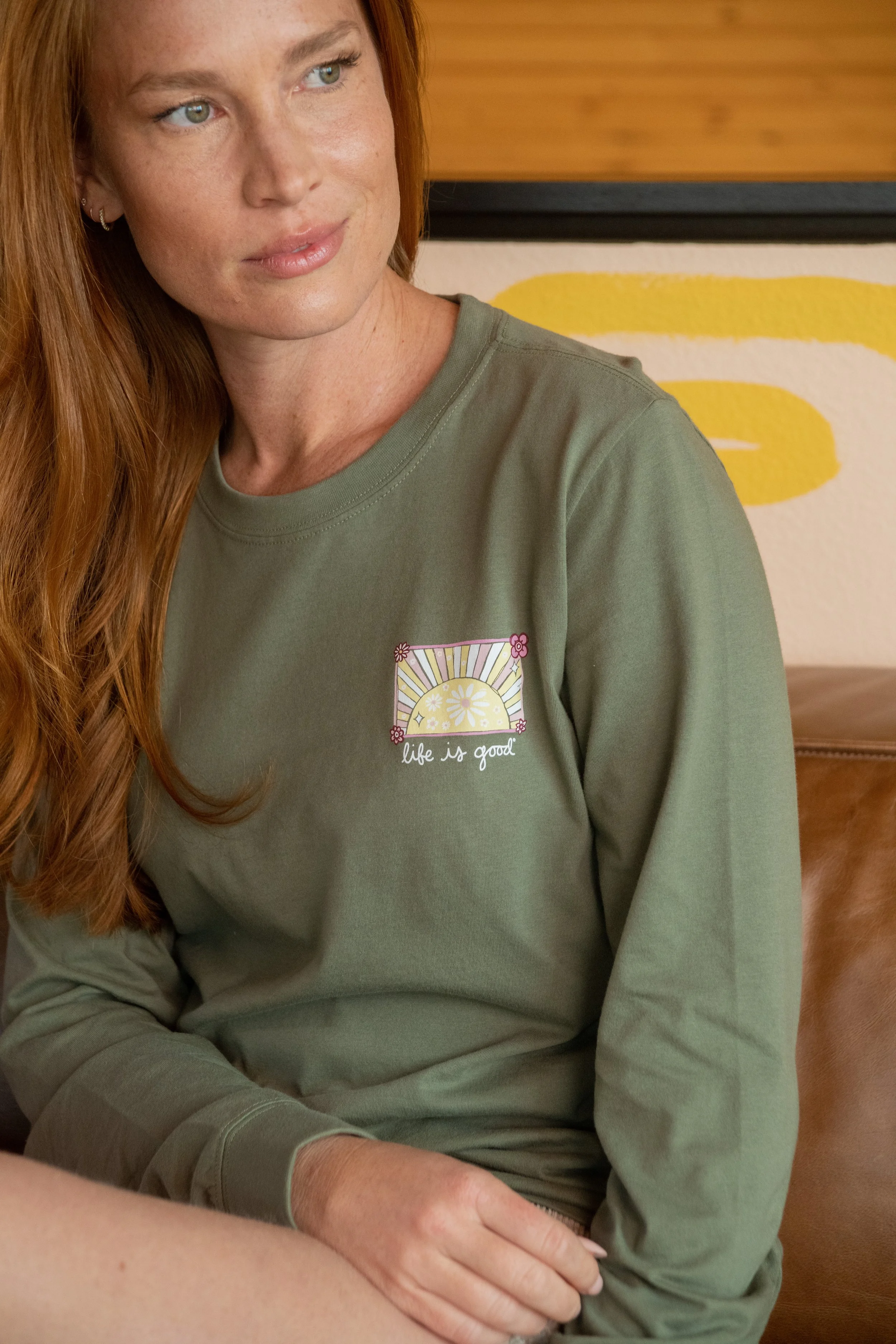 A woman with red hair and freckles wearing an olive green sweatshirt with a graphic of a sun and the phrase 'life is good' on it.