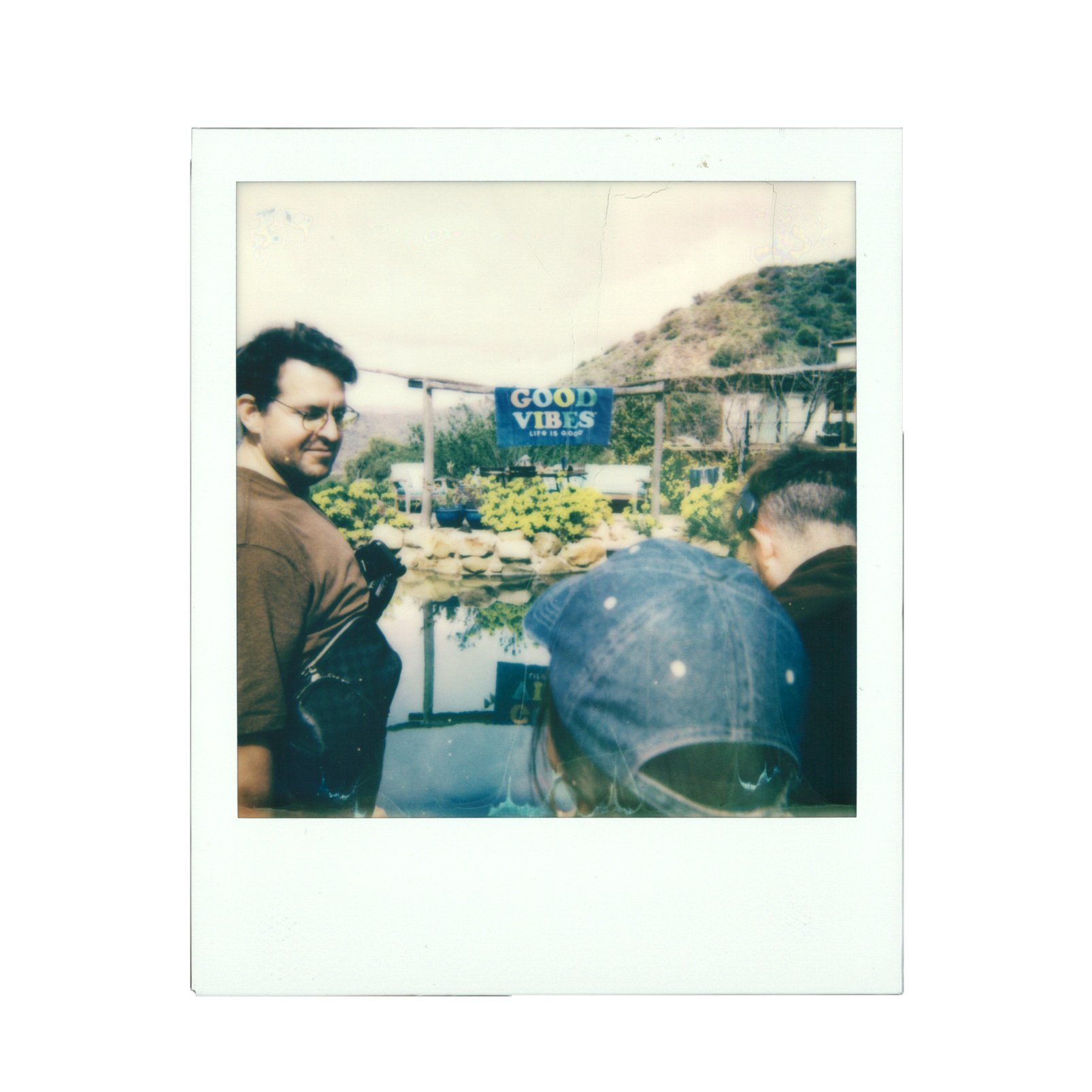 A vintage Polaroid photo showing people near a pond with a sign that says "Good Vibes" and a hillside in the background.