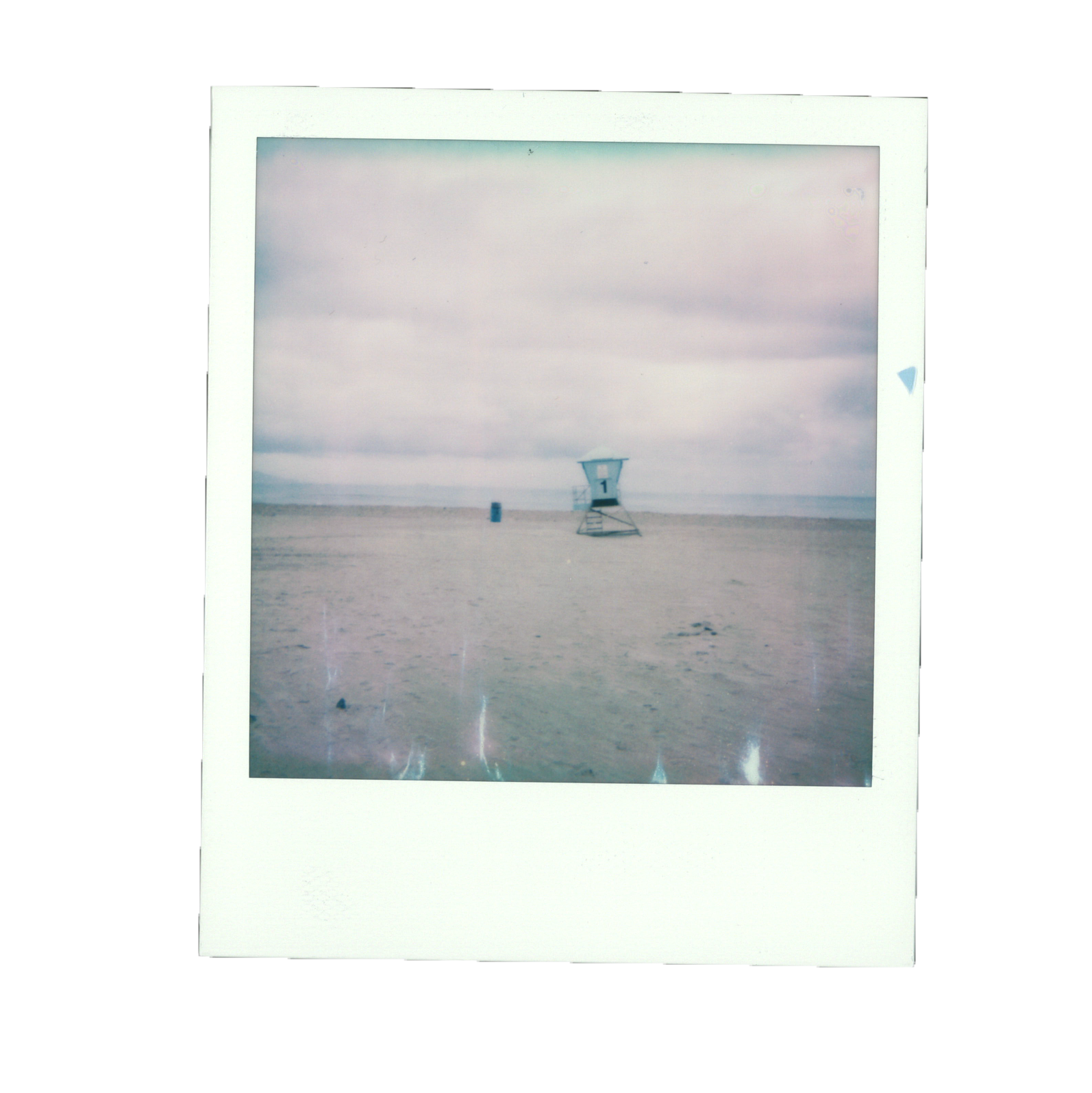 A vintage Polaroid photo of a lifeguard tower on a sandy beach under cloudy skies, with a blue trash can nearby.