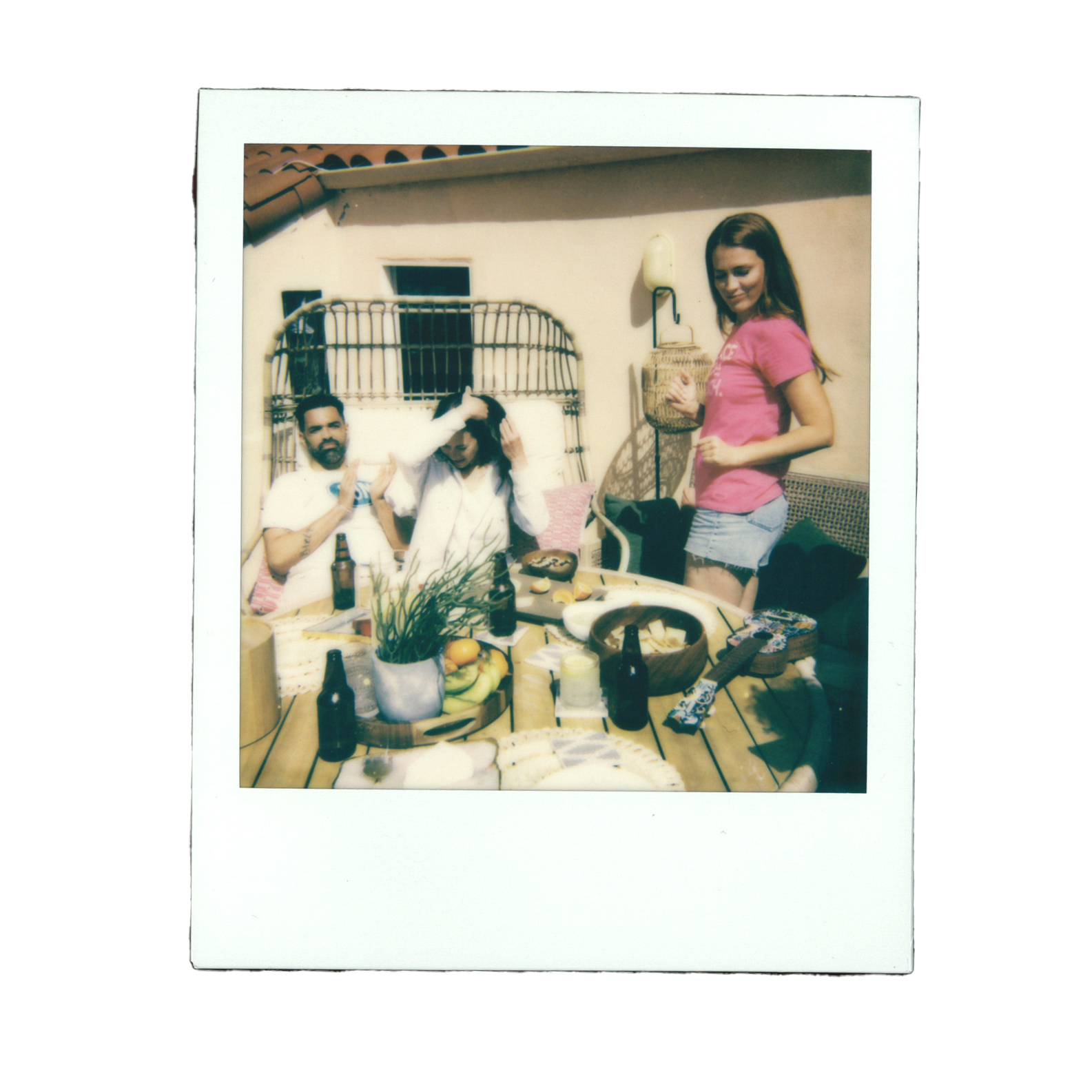 Three people at a patio table with food and drinks. Two seated, one standing. Bright outdoor setting with a beige wall and window.