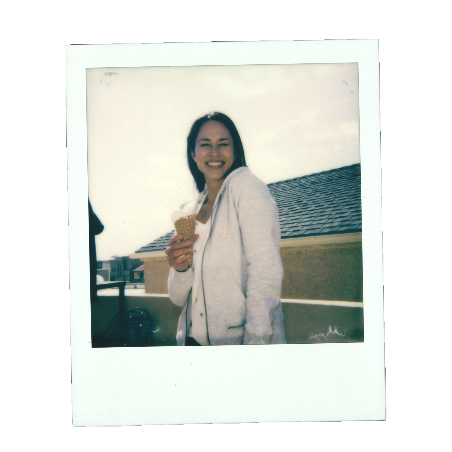 A woman smiling and holding an ice cream cone on a rooftop balcony with a view of neighboring buildings and a cloudy sky.