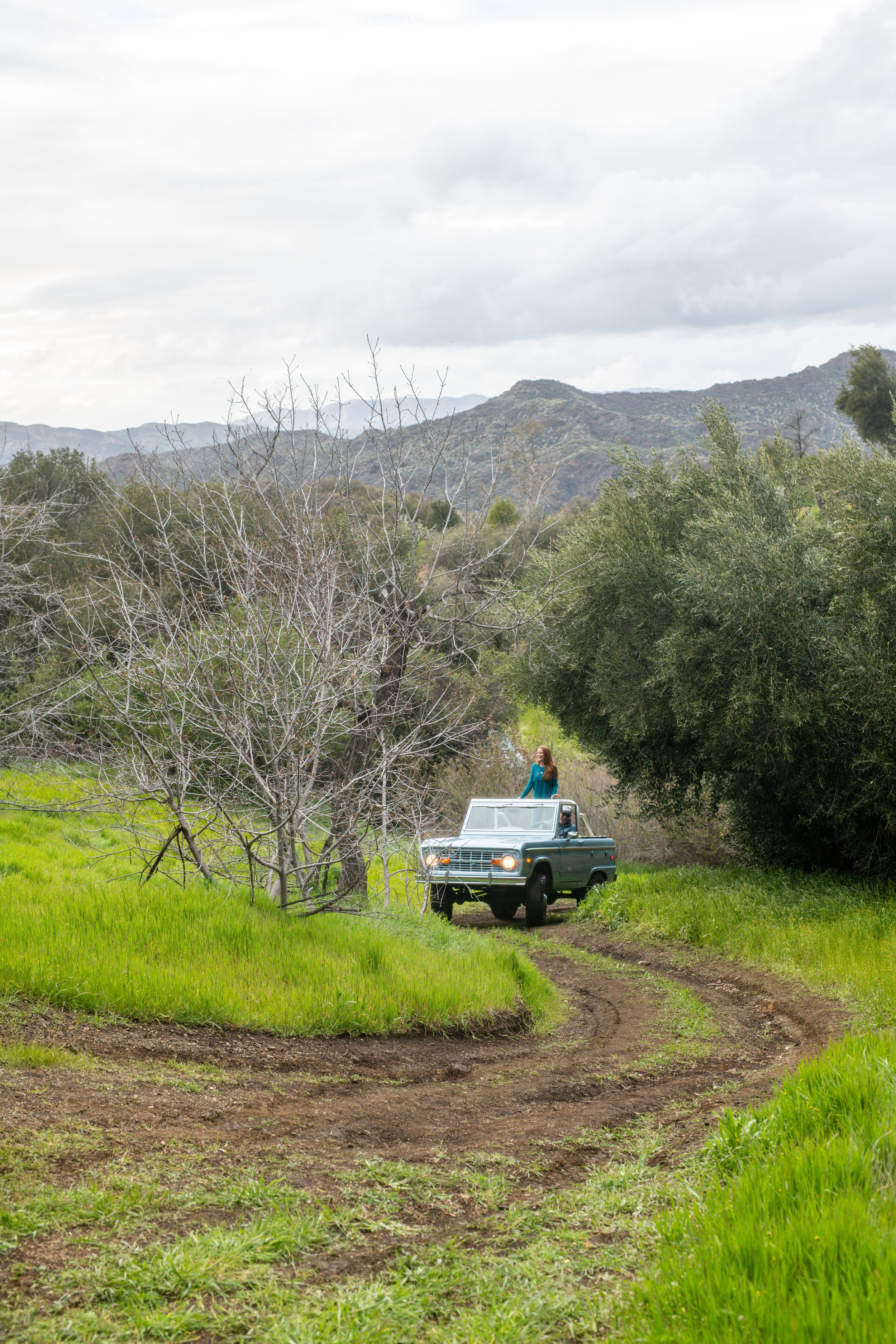 A woman in a blue dress standing in the back of a vintage blue pickup truck on a dirt path surrounded by green grass and trees, with mountains and cloudy sky in the background.