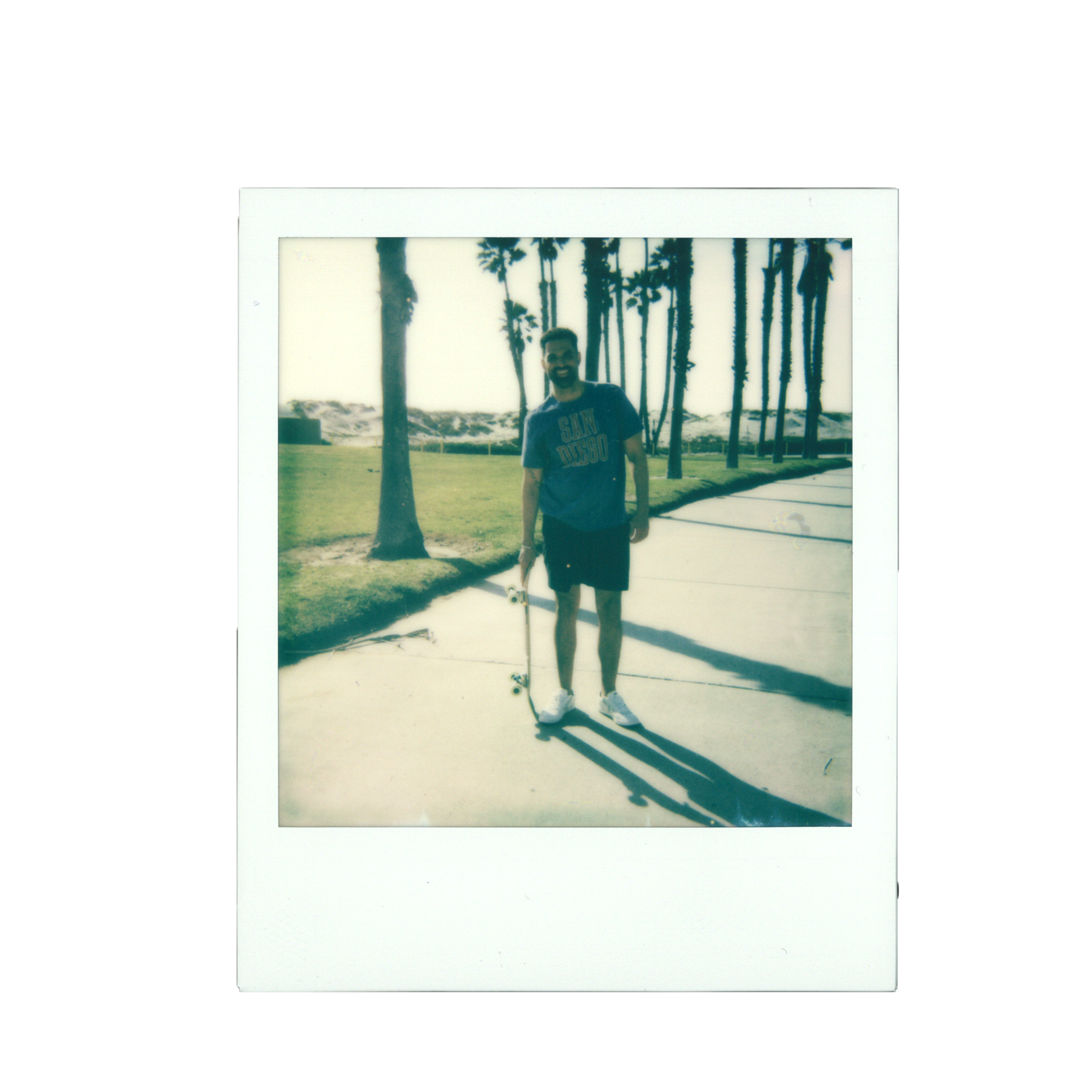 A man standing on a concrete pathway holding a skateboard at a beachside park, with palm trees and sand dunes in the background, under a bright sky.
