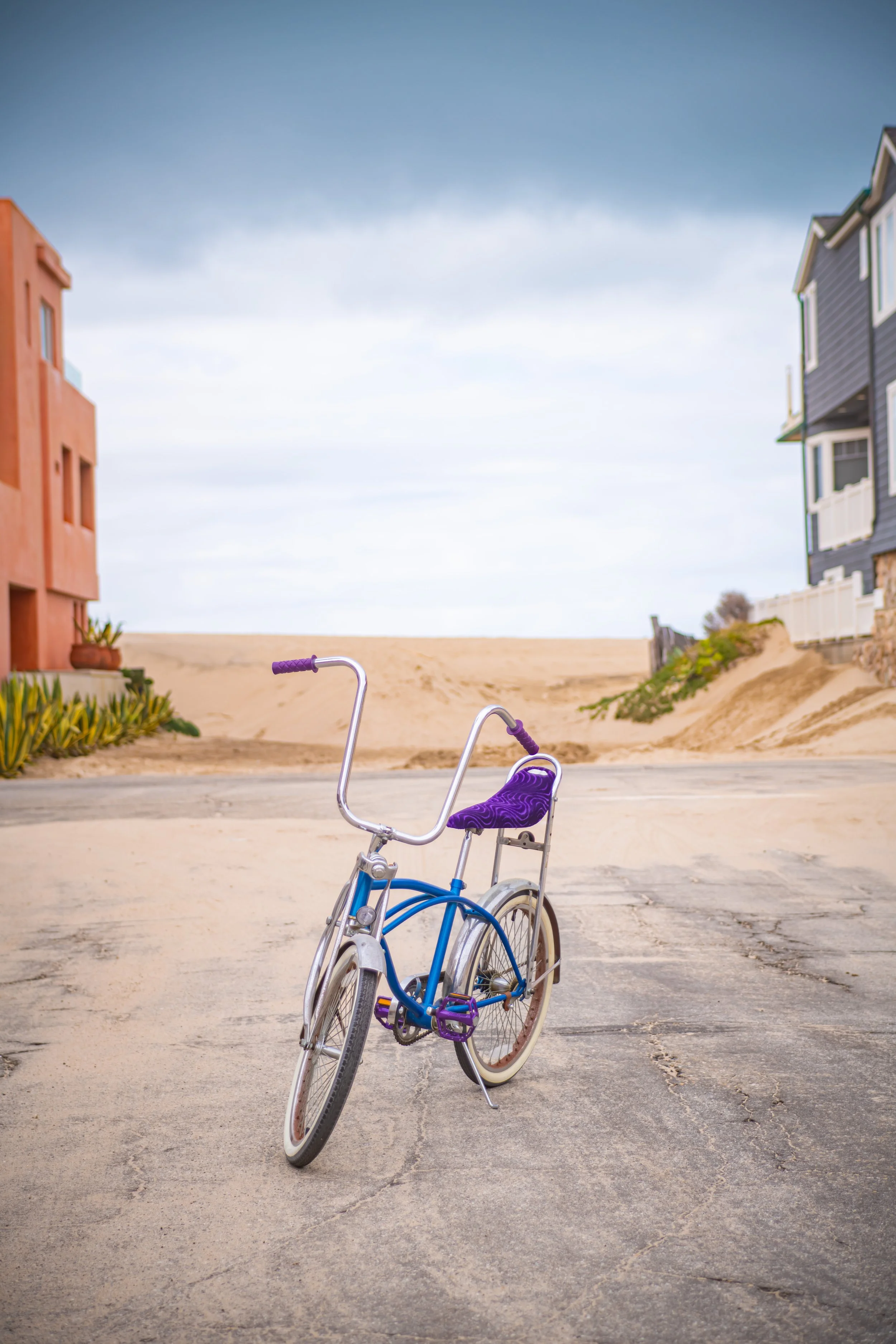 A vintage purple and blue bicycle parked on a paved beach area under a cloudy sky, with sand dunes and colorful houses in the background.