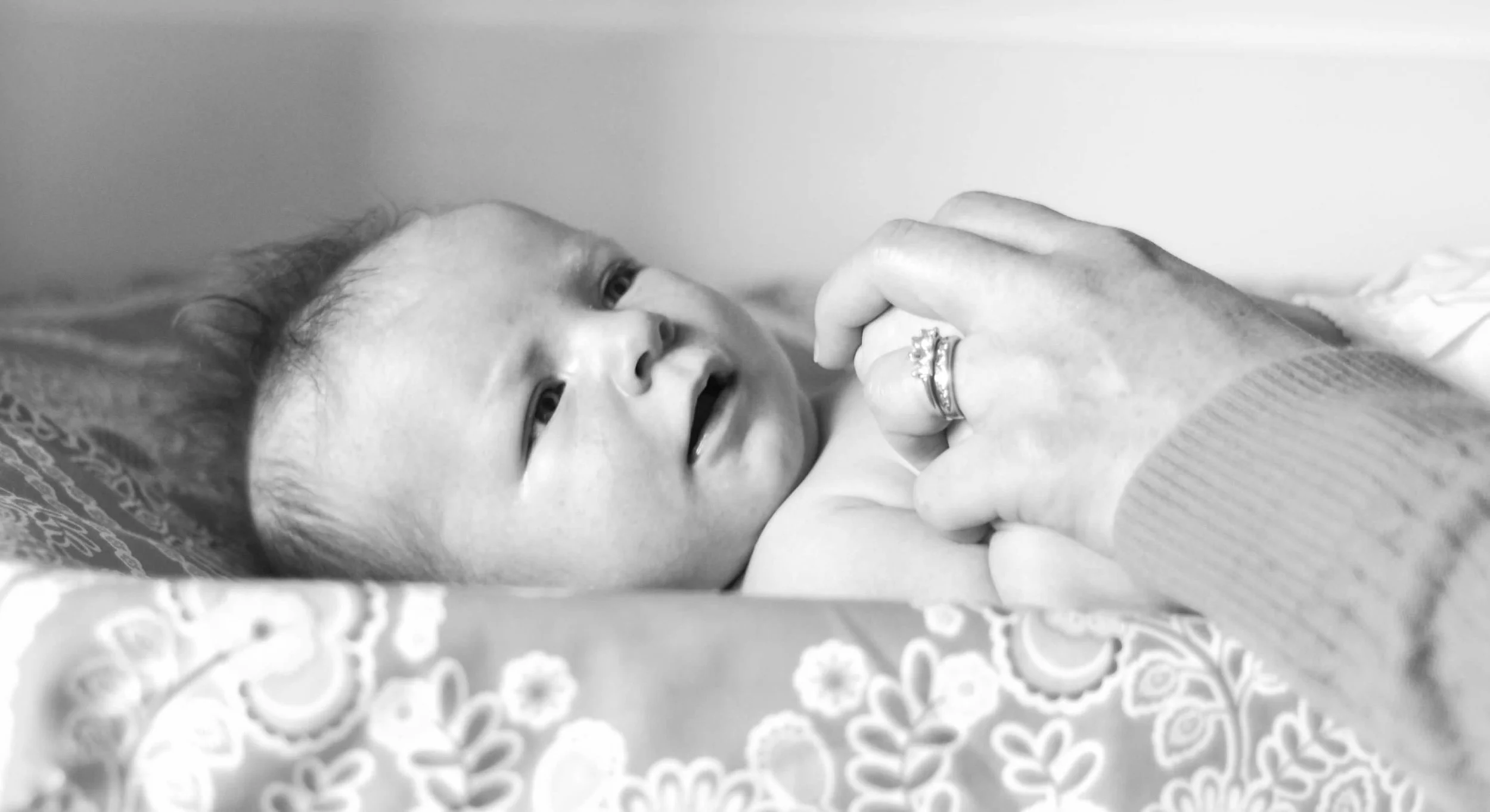 A baby lying on a bed, looking up, with an adult's hand gently holding the baby's hand. The adult is wearing rings on their fingers, and the baby appears to be smiling or speaking.