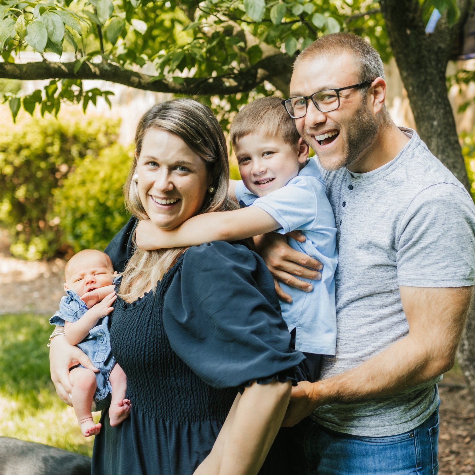 A smiling family of four poses outdoors under a tree. The mother holds a baby, while the father and a young child embrace her. They are dressed casually, enjoying a sunny day in a garden setting.