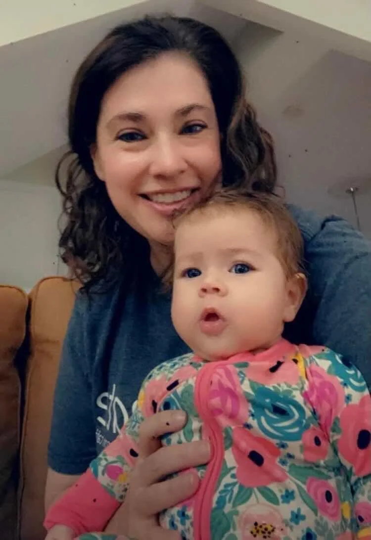 A woman with dark wavy hair smiling and holding a baby girl with curly hair and blue eyes. The woman is wearing a blue shirt, and the baby is dressed in a colorful floral-patterned jacket.