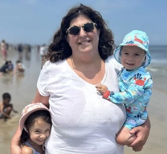 A woman wearing sunglasses and a white shirt holding two children, one girl in a striped outfit and pink hat, and one baby in a colorful sun hat and outfit, standing on a crowded beach.
