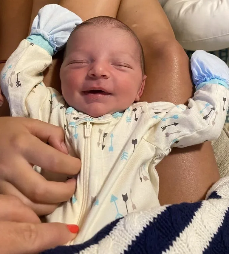 Smiling baby lying on an adult's lap, wearing a white onesie with arrow patterns, with hands in blue mittens, being gently held and supported.