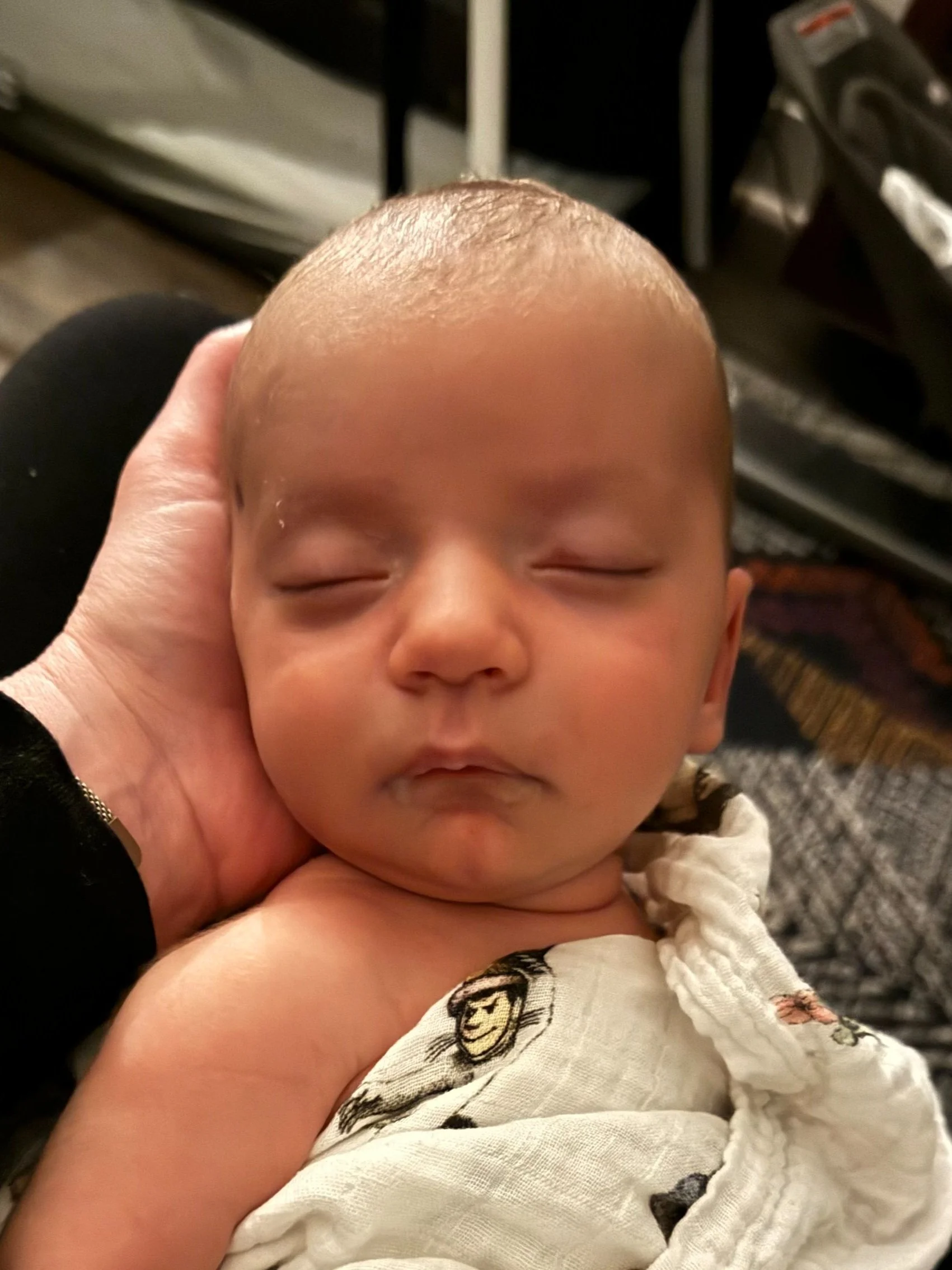 A newborn baby sleeping peacefully with a hand supporting its head, wrapped in a white blanket with a monkey print.