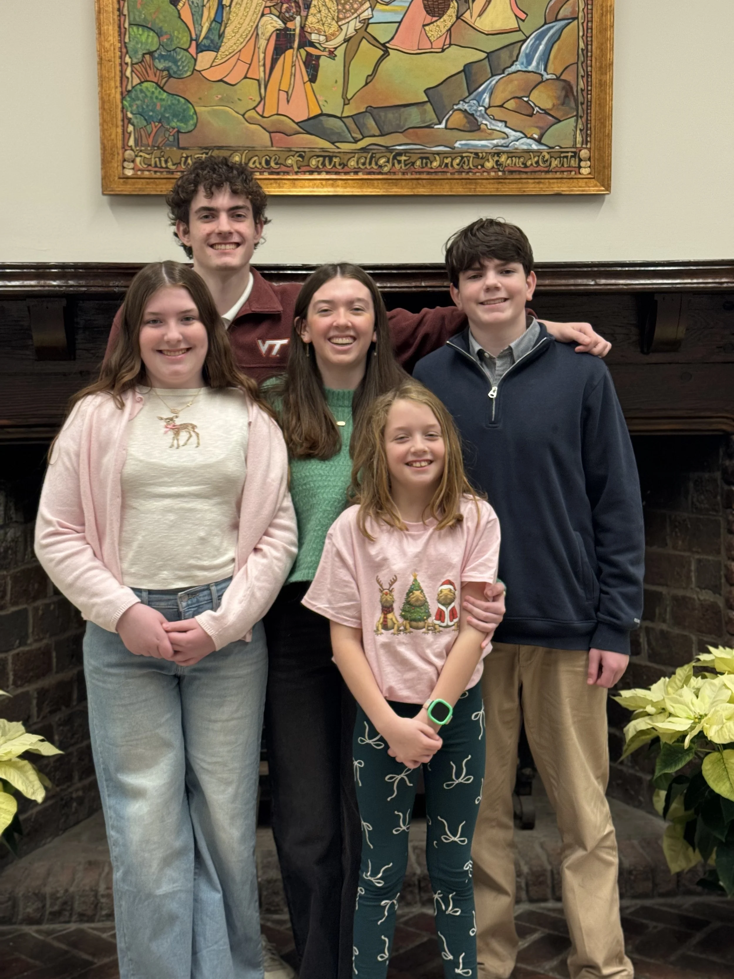 A group of six young people, four females and two males, standing together indoors in front of a brick fireplace with a painting above it. They are smiling and appear to be enjoying each other's company.