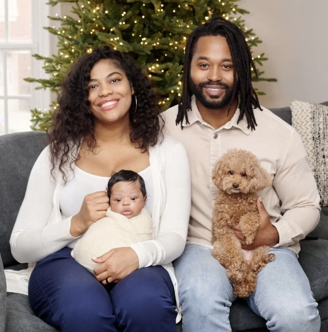 Family with baby and dog sitting on couch beside Christmas tree.