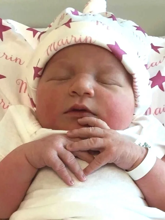 Newborn baby wearing a white outfit and a hat with pink stars and text, resting with hands clasped over chest.