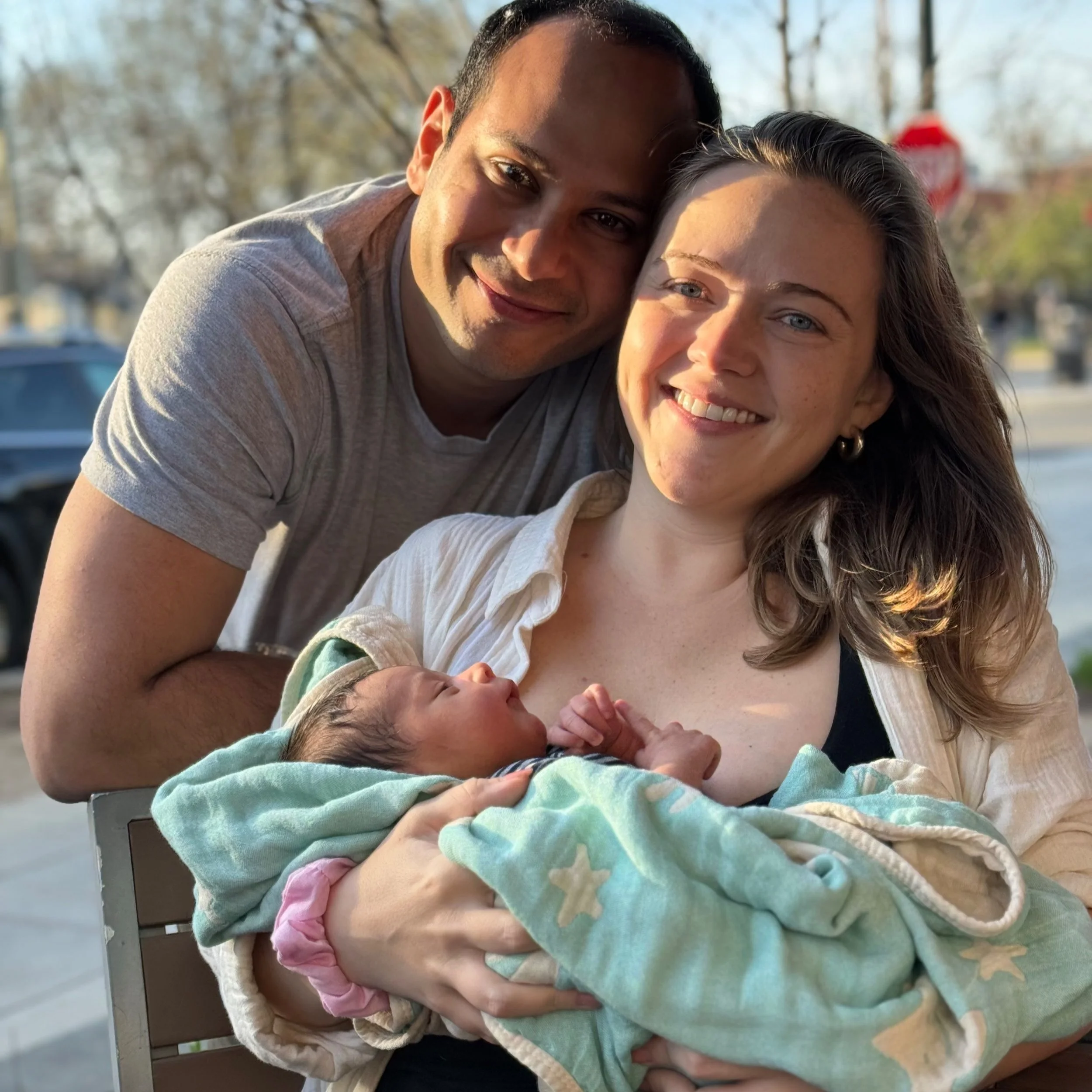 A smiling couple holding a sleeping baby wrapped in a green blanket with star patterns, outdoors with trees in the background.
