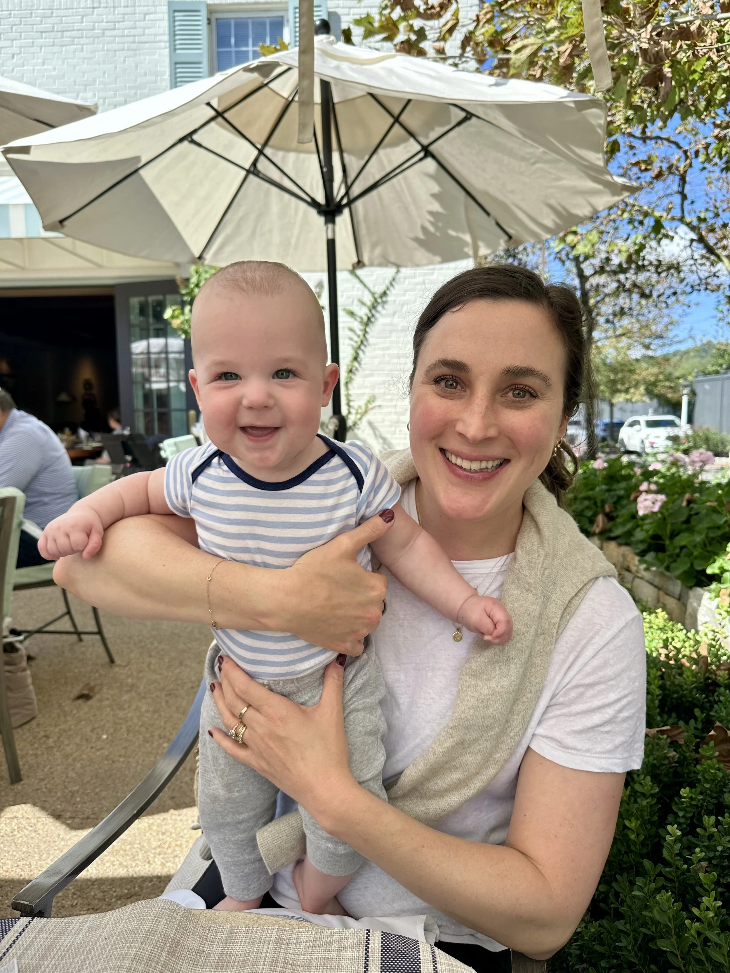 A smiling woman holding a happy baby outside at a restaurant or cafe, with a large beige umbrella and greenery in the background.