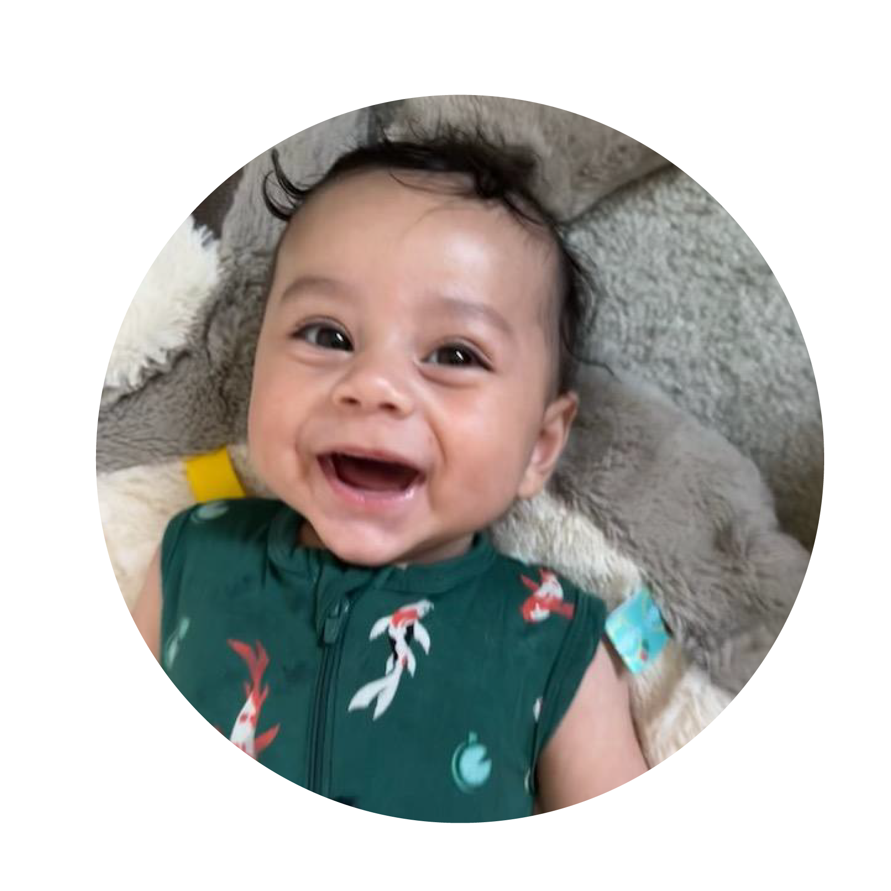 A happy baby wearing a green shirt with a fish pattern, smiling while lying on a soft cushion.