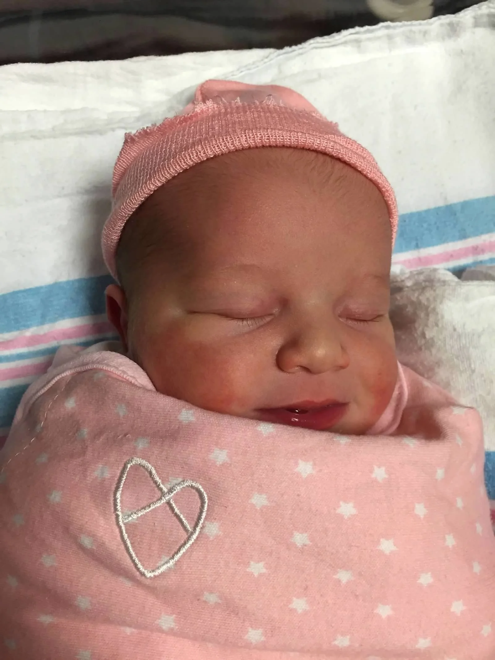 Newborn baby swaddled in a pink blanket with a hat, sleeping on a hospital blanket.