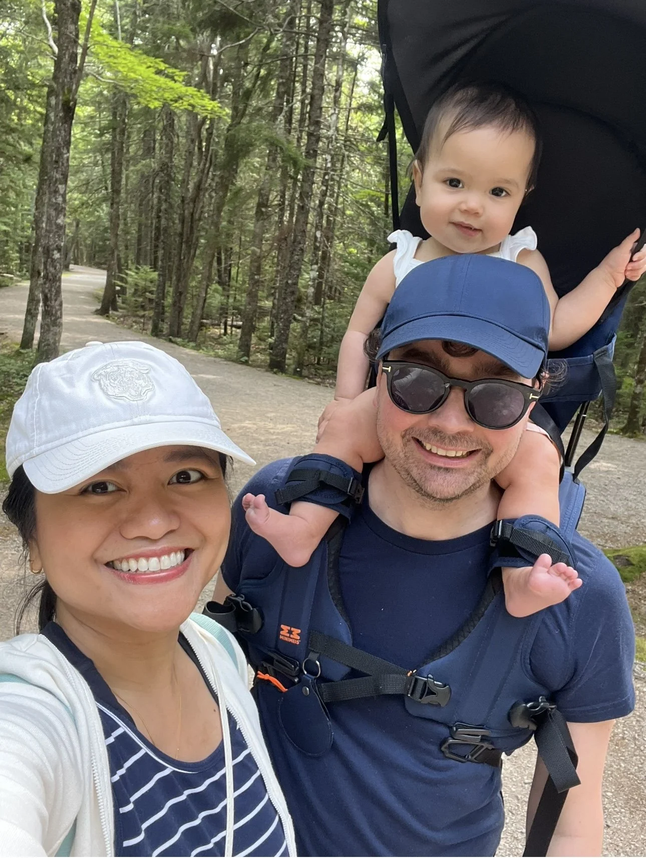 A couple with a baby on a hiking trail in a forest. The man is wearing sunglasses and has the baby in a backpack carrier. The woman is smiling and wearing a cap.