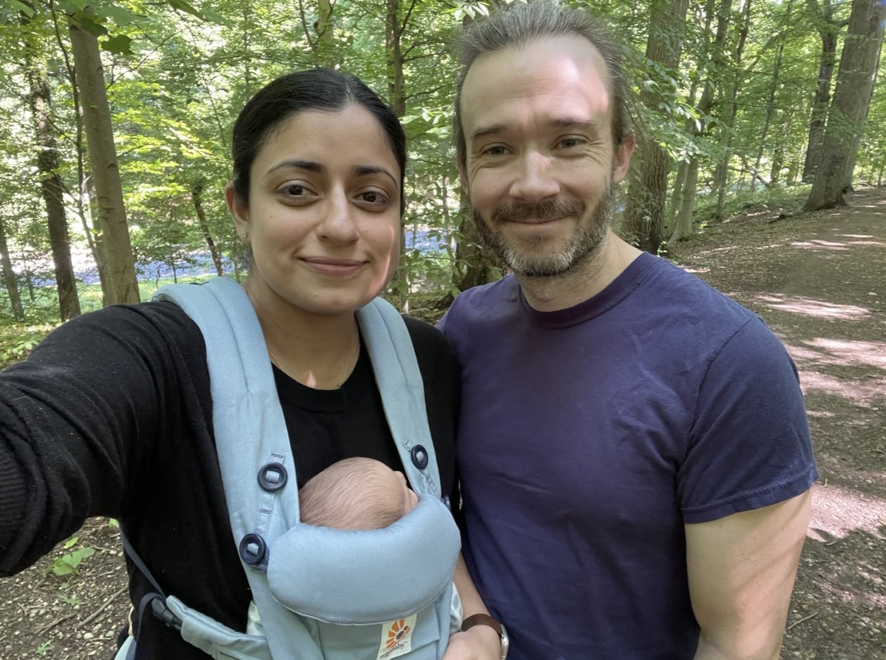 A couple with a baby in a carrier walking in a wooded area.